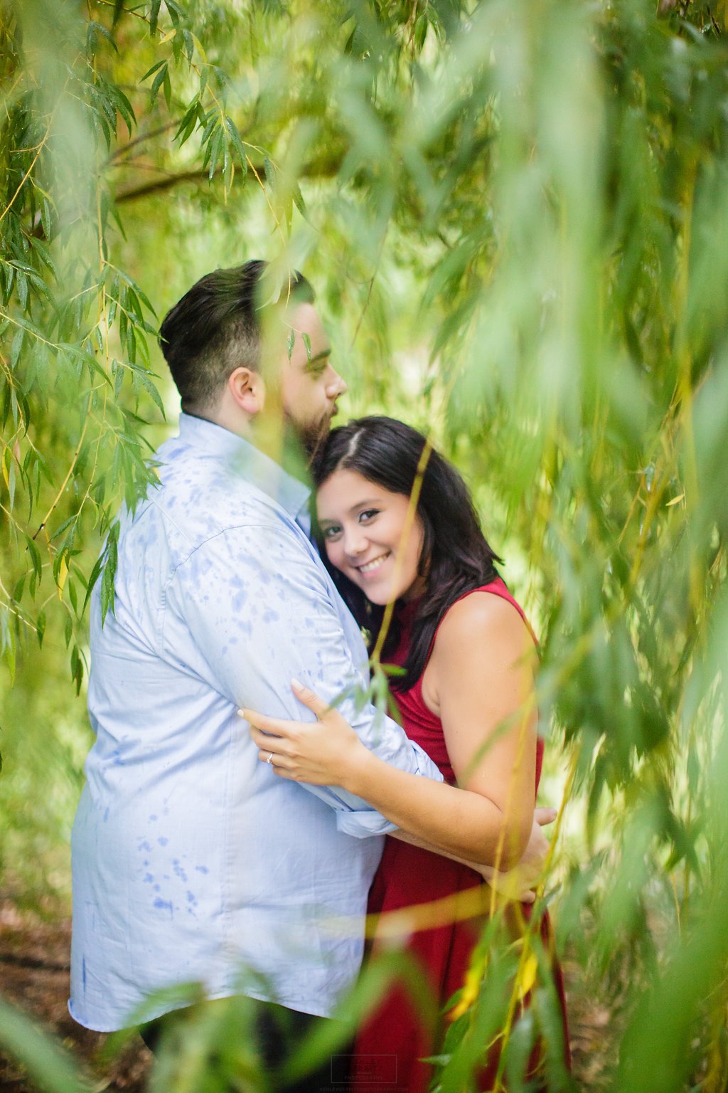 A young couple embraces in a lush, green outdoor setting surrounded by hanging branches and leaves, with the woman smiling at the camera.