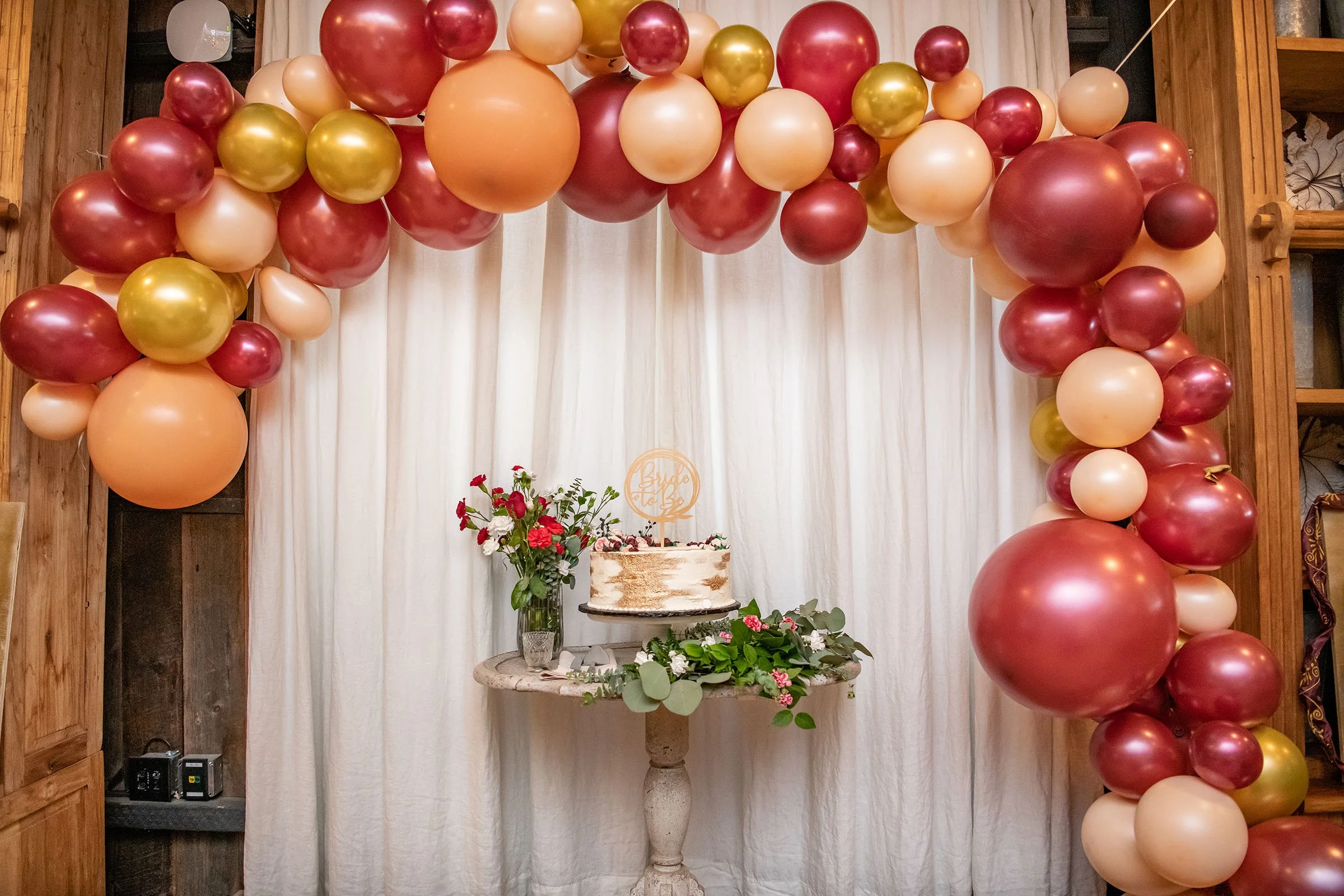 Decorative balloon arch with maroon, gold, and cream balloons framing a white curtain backdrop, with a table featuring a cake, pink flowers, and greenery.