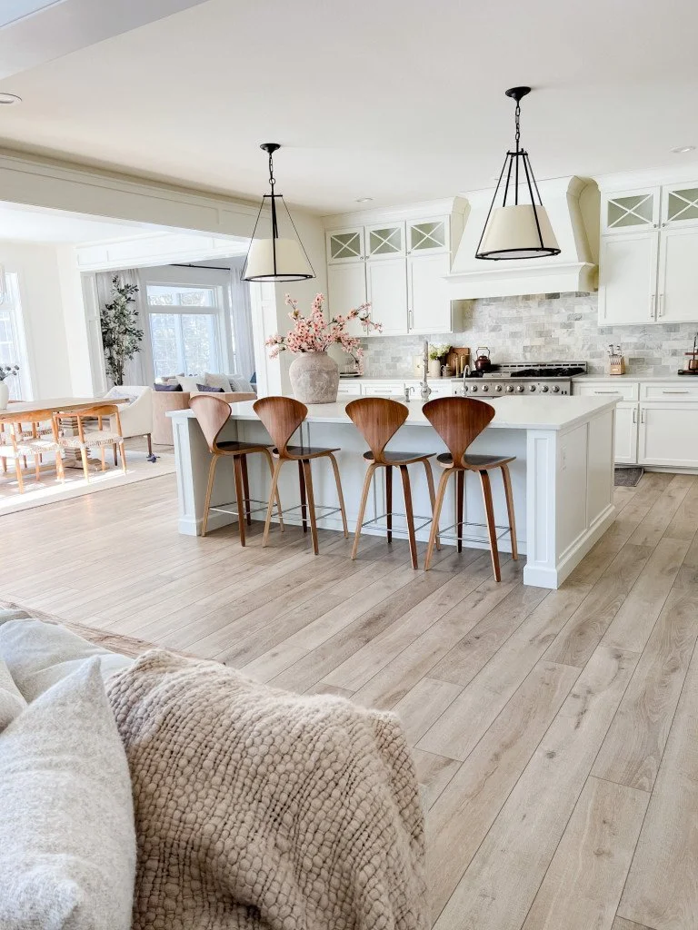 Modern kitchen with white cabinetry, island with four wooden bar stools, pink flower arrangement in a large vase, stainless steel stove, light wood flooring, and adjacent dining and living areas.