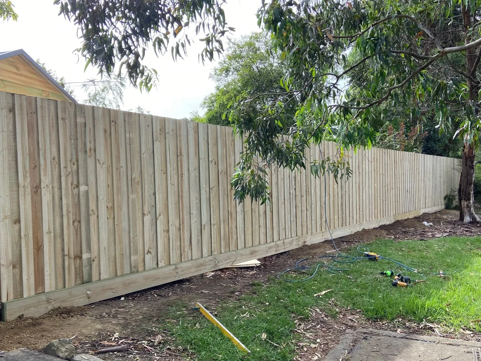 Wooden fence under construction next to a tree with tools and wires on the ground.