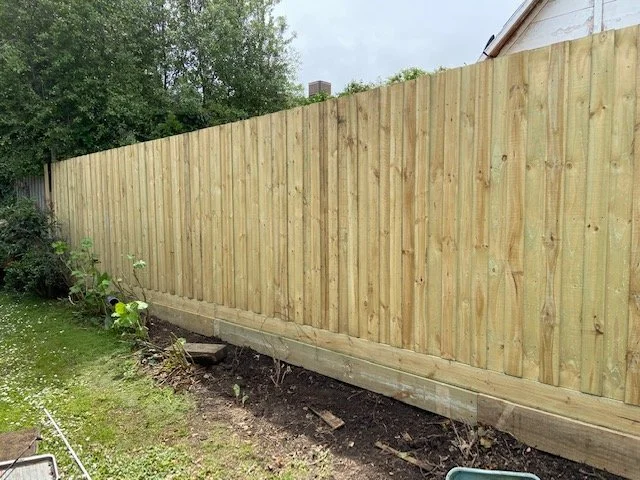 Newly installed wooden privacy fence with vertical planks, supported by a concrete base, in a backyard with a small garden bed and green grass.