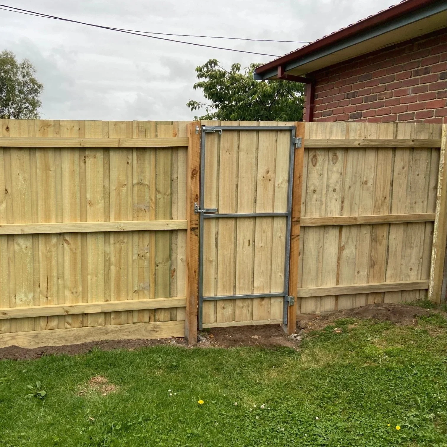A wooden privacy fence with a metal framed gate on a grassy yard with patches of soil, and a red brick house partially visible in the background under a cloudy sky.