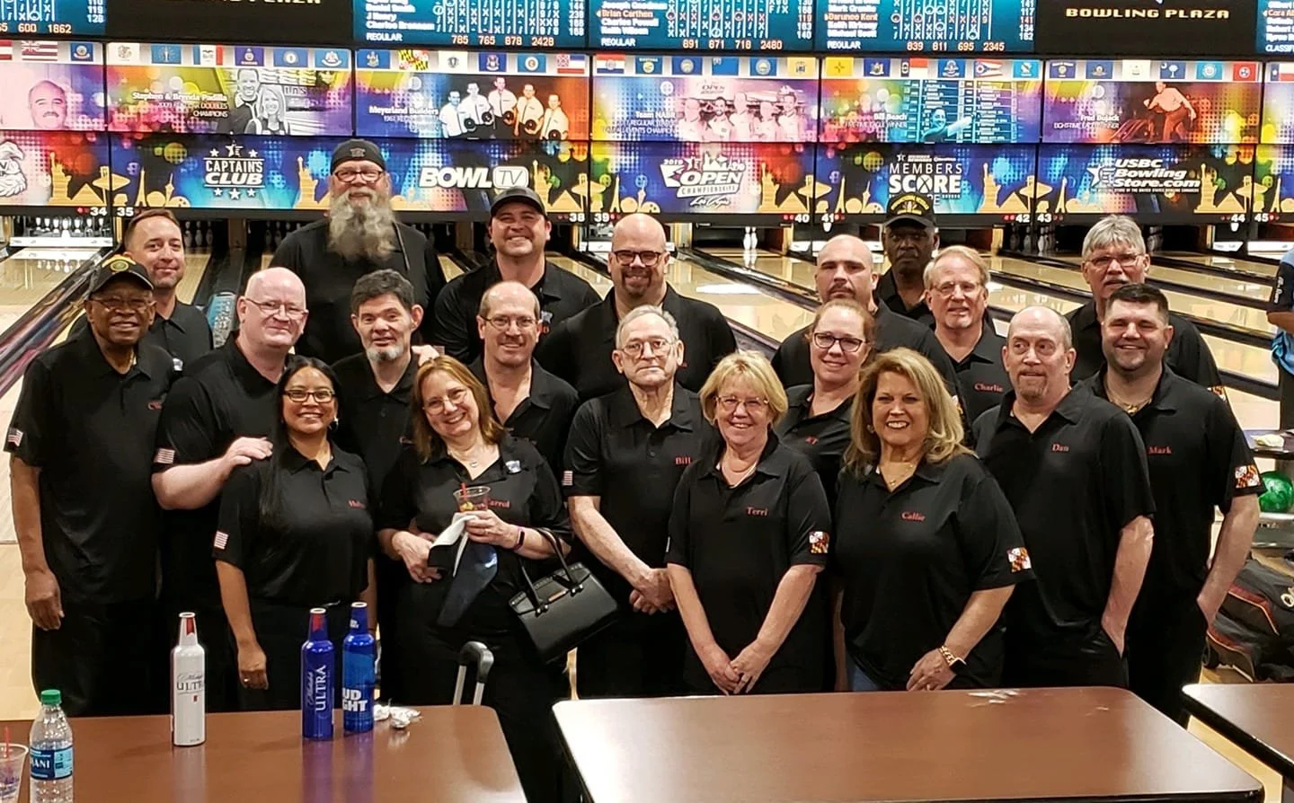 Scott Gielda's Nationals Team of people in black shirts posing for a photo at a bowling alley, with digital scoreboards and bowling lanes in the background.