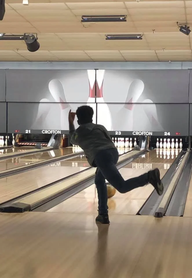 A person in a gray hoodie and jeans is in the act of bowling, releasing a yellow bowling ball down a lane at a bowling alley. They are captured mid-throw with one leg lifted behind them.
