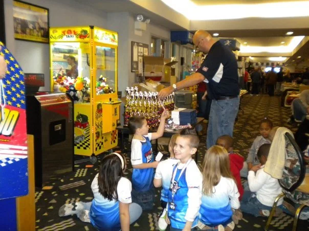 Children waiting to get prizes from a Coach Scott at a gaming booth in an arcade or gaming area.