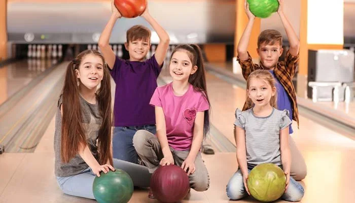 Five children in a bowling alley, three girls and two boys, sitting on the floor with bowling balls, two boys holding bowling balls above their heads.
