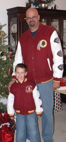 Scott Gielda and a young Markie standing in front of a Christmas tree, smiling, both wearing Washington Redskins jackets. The man is bald with glasses, and the boy has short hair.