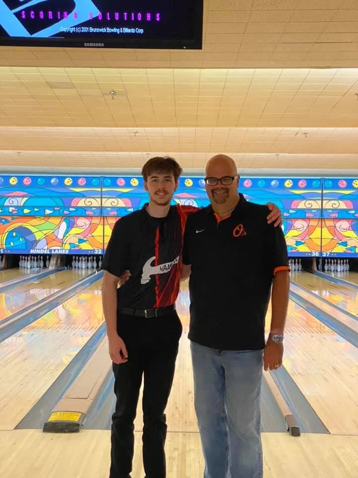 Markie and Scott standing together at a bowling alley, smiling, with bowling lanes and colorful lights in the background.