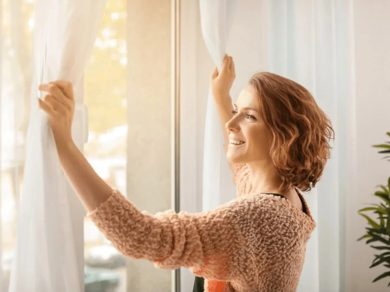 A woman with curly red hair smiling and pulling back white curtains in a bright room with large windows and a potted plant.