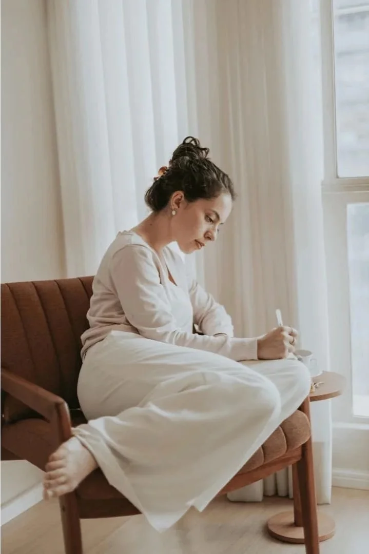A woman sitting on a brown chair near a window, wearing light-colored clothing, writing in a notebook with a pen.