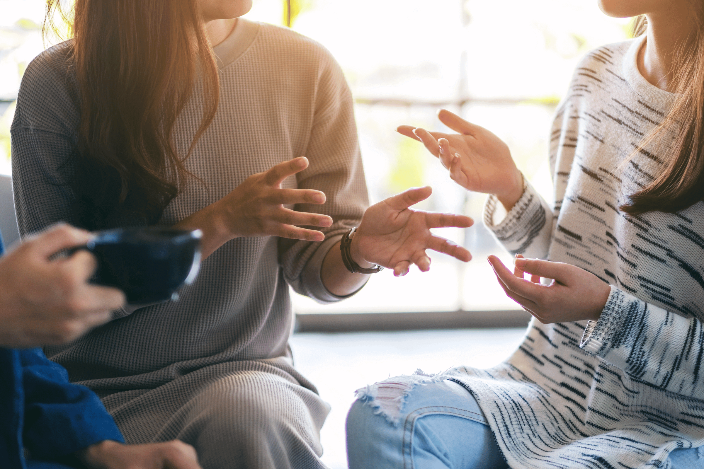 Two women are engaged in a conversation, using hand gestures, with one holding a mug, in a well-lit indoor setting.