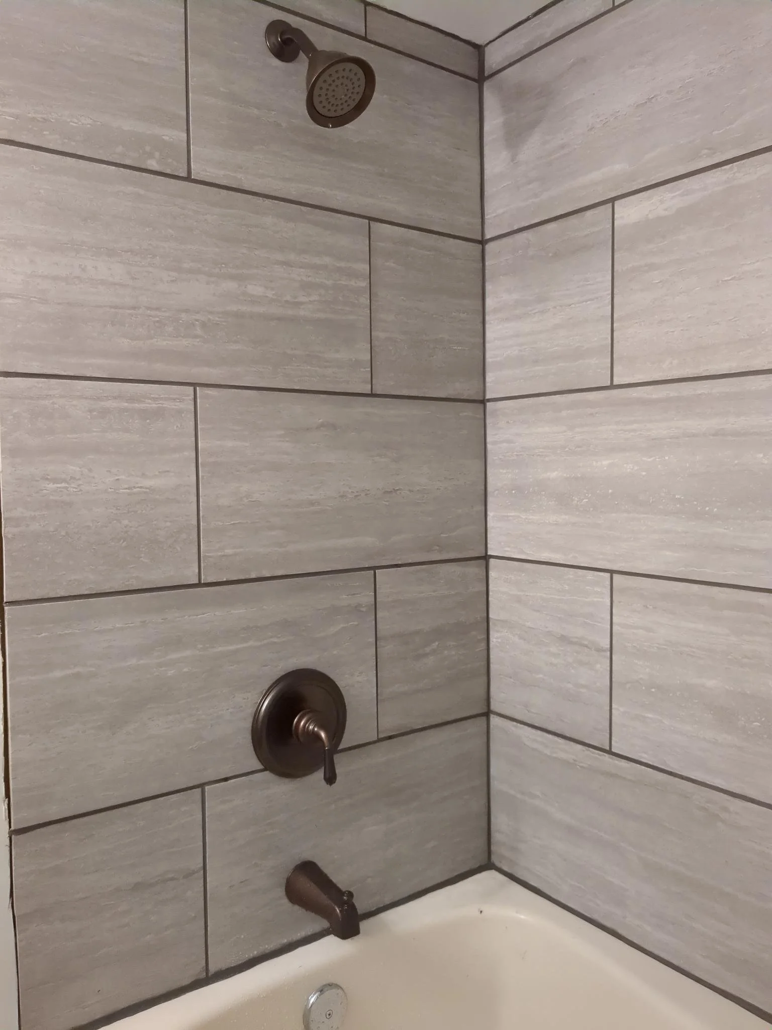 Bathroom shower with gray rectangular tiles, a wall-mounted showerhead, a round hot/cold water control valve, and a faucet spout over a white bathtub.