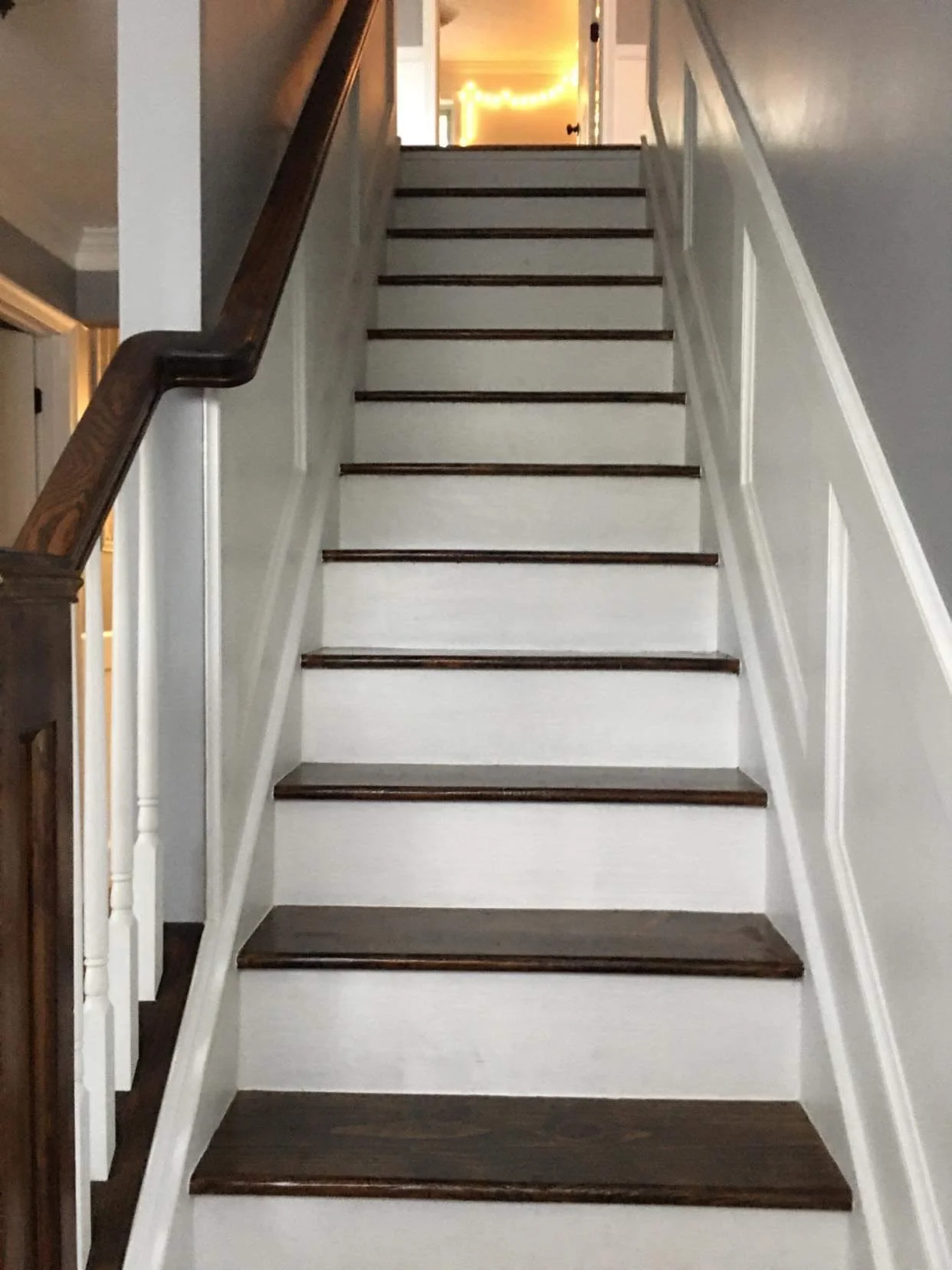Wooden staircase with dark brown steps, white risers, and white walls, leading up to a doorway with warm lighting.