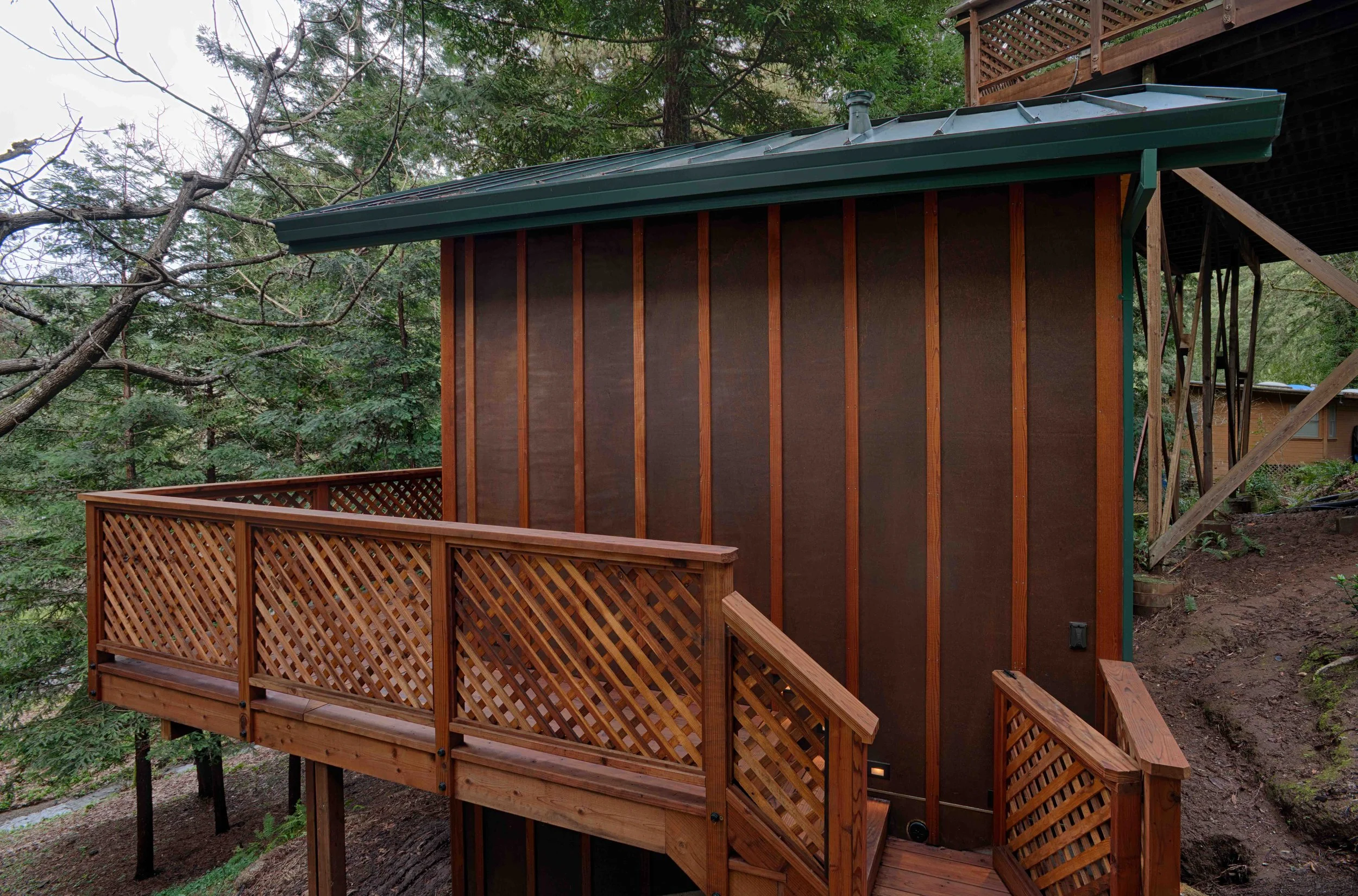 Side view of a wooden house with a small deck surrounded by trees in a wooded area.