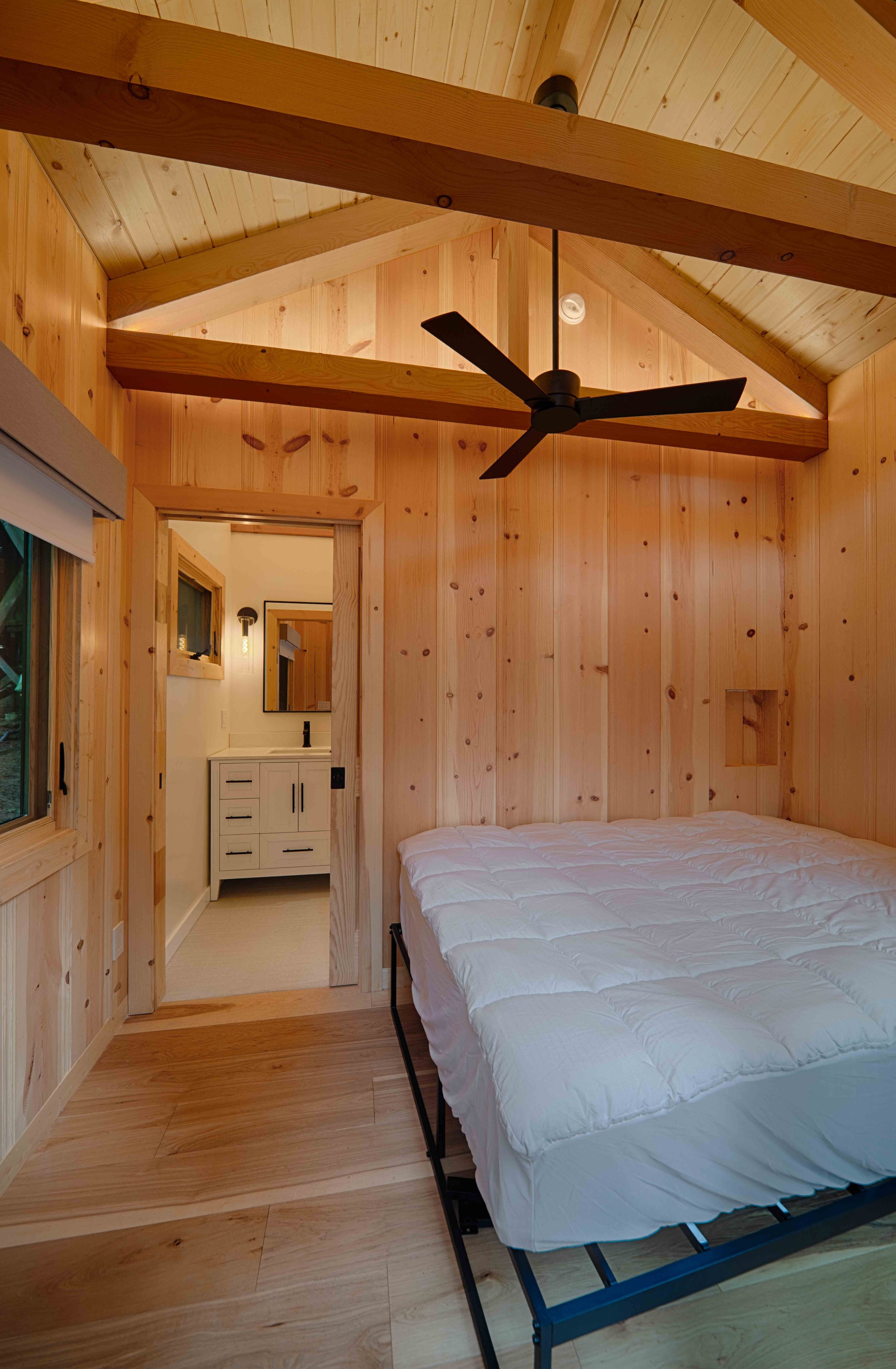 A cozy bedroom with wood-paneled walls and ceiling, a black ceiling fan, a bed with a white quilt, and a window on the left. In the background, there is a doorway leading to a bathroom with a white vanity and a mirror.