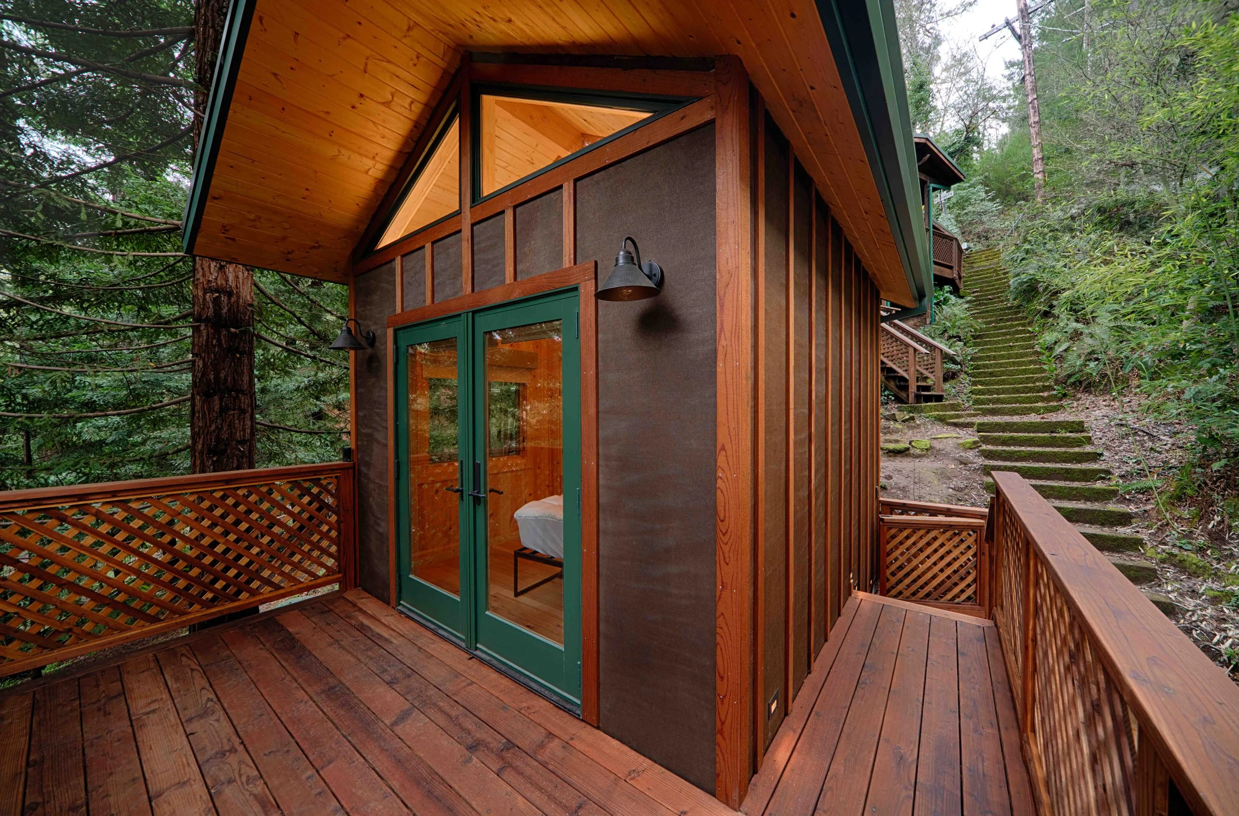 A small wooden cabin with green door and windows, surrounded by trees and a forested hillside with steps leading up