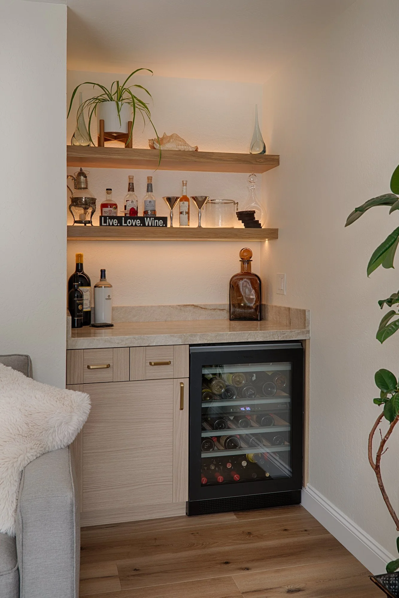 Home bar with wooden shelves holding liquor bottles, glasses, and decorative items. A wine cooler is built into the cabinet below the countertop, which has a light-colored wood finish. There are also some bottles on the countertop and a sign that reads "Live. Love. Wine." The setup includes potted plants and decorative glassware.