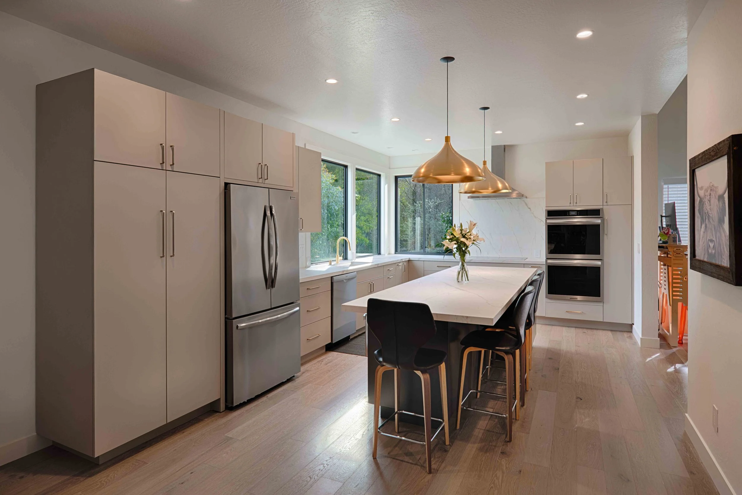 Modern kitchen with light-colored cabinets, stainless steel refrigerator, white marble island with black and tan chairs, large windows, gold pendant lights, and built-in double oven.
