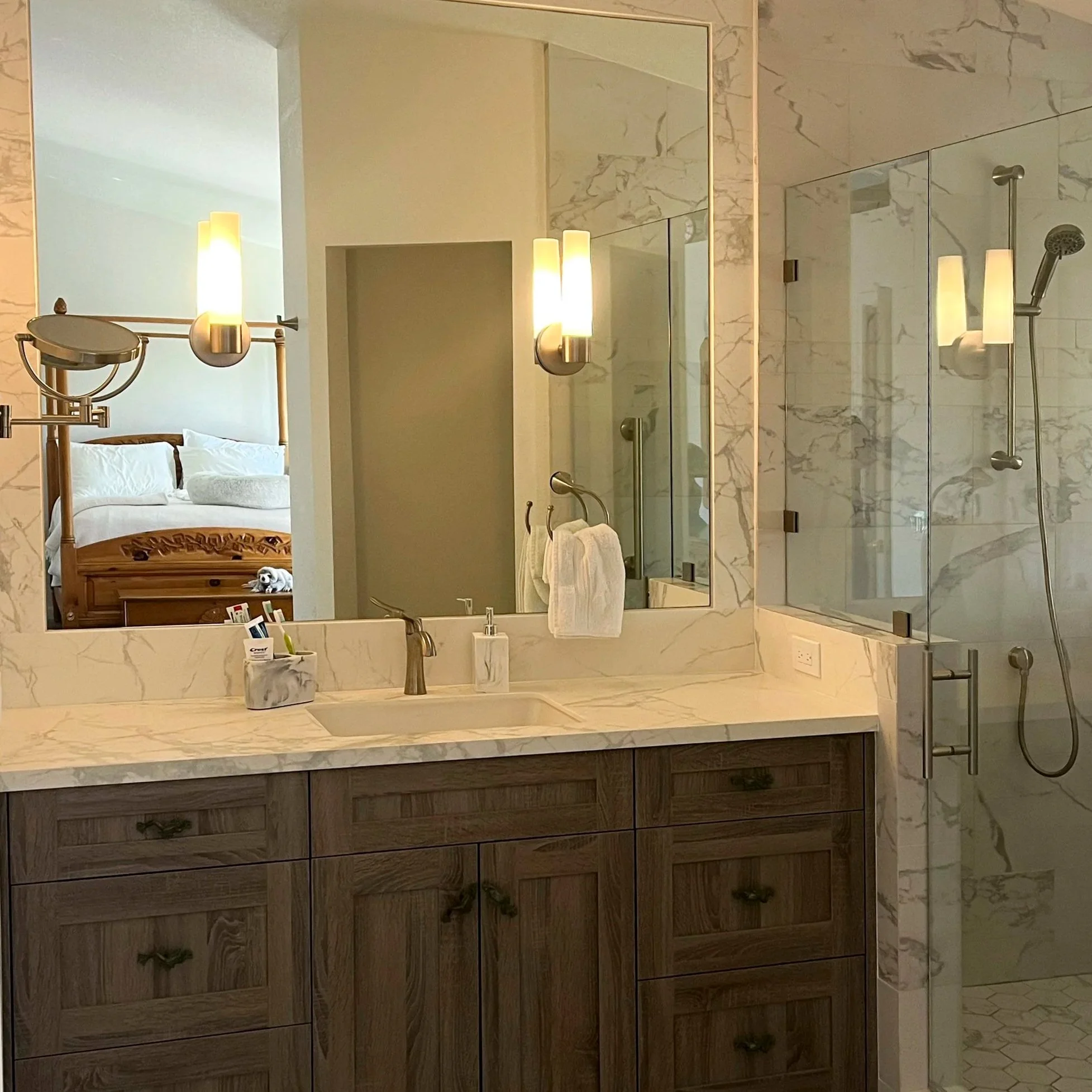 A modern bathroom with a marble countertop, a sink with a bronze faucet, wooden cabinets, two wall-mounted light fixtures, a large mirror, a glass-enclosed shower with marble walls, a showerhead, a soap dispenser, and a towel hanging on a hook. Beyond the bathroom, a bedroom with a wooden bed, white linens, pillows, and a nightstand is visible.