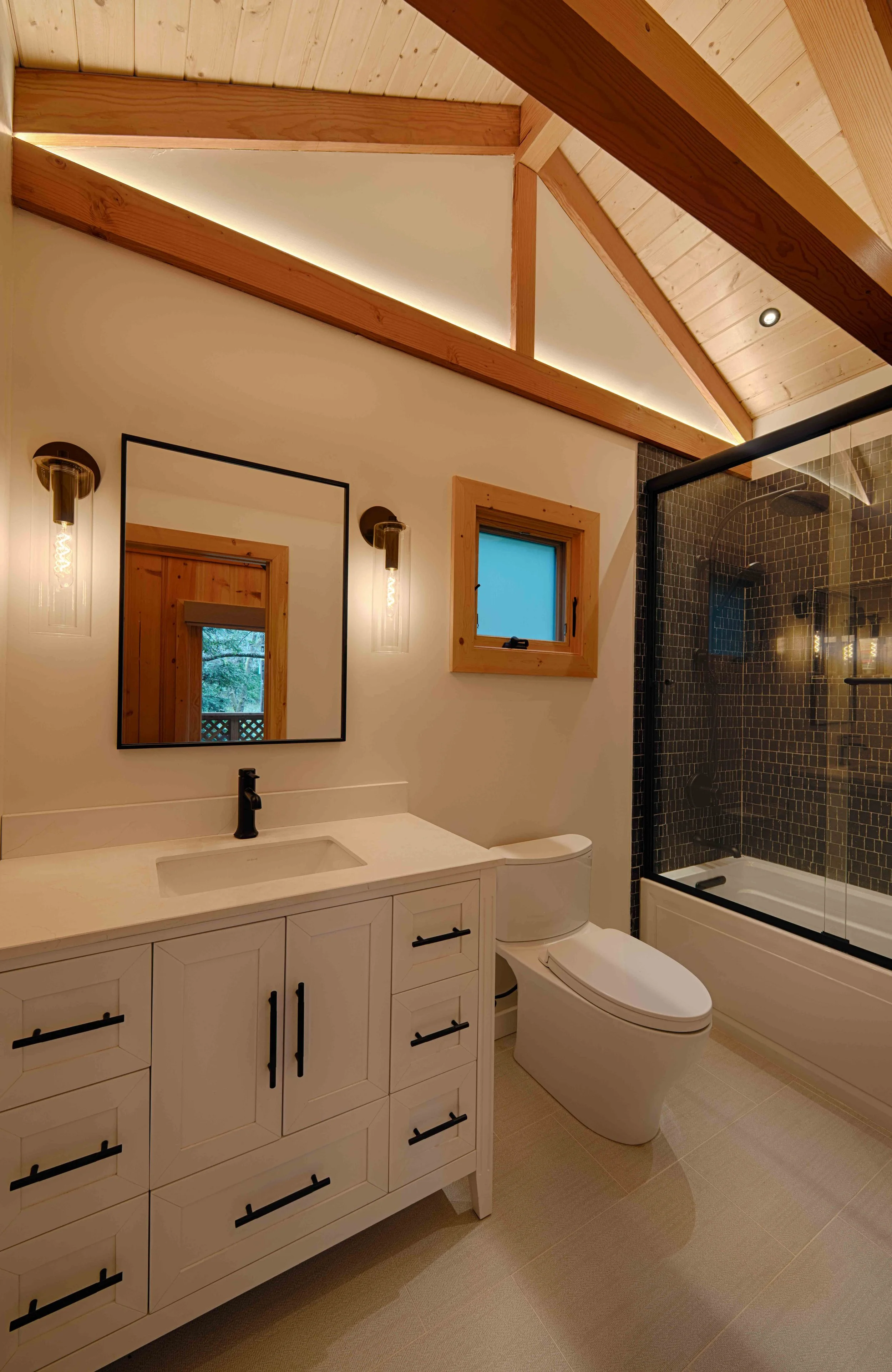 Bathroom with wooden ceiling beams, a white vanity with a black faucet, a large mirror, a window, a modern toilet, and a shower with dark mosaic tiles and a glass enclosure.
