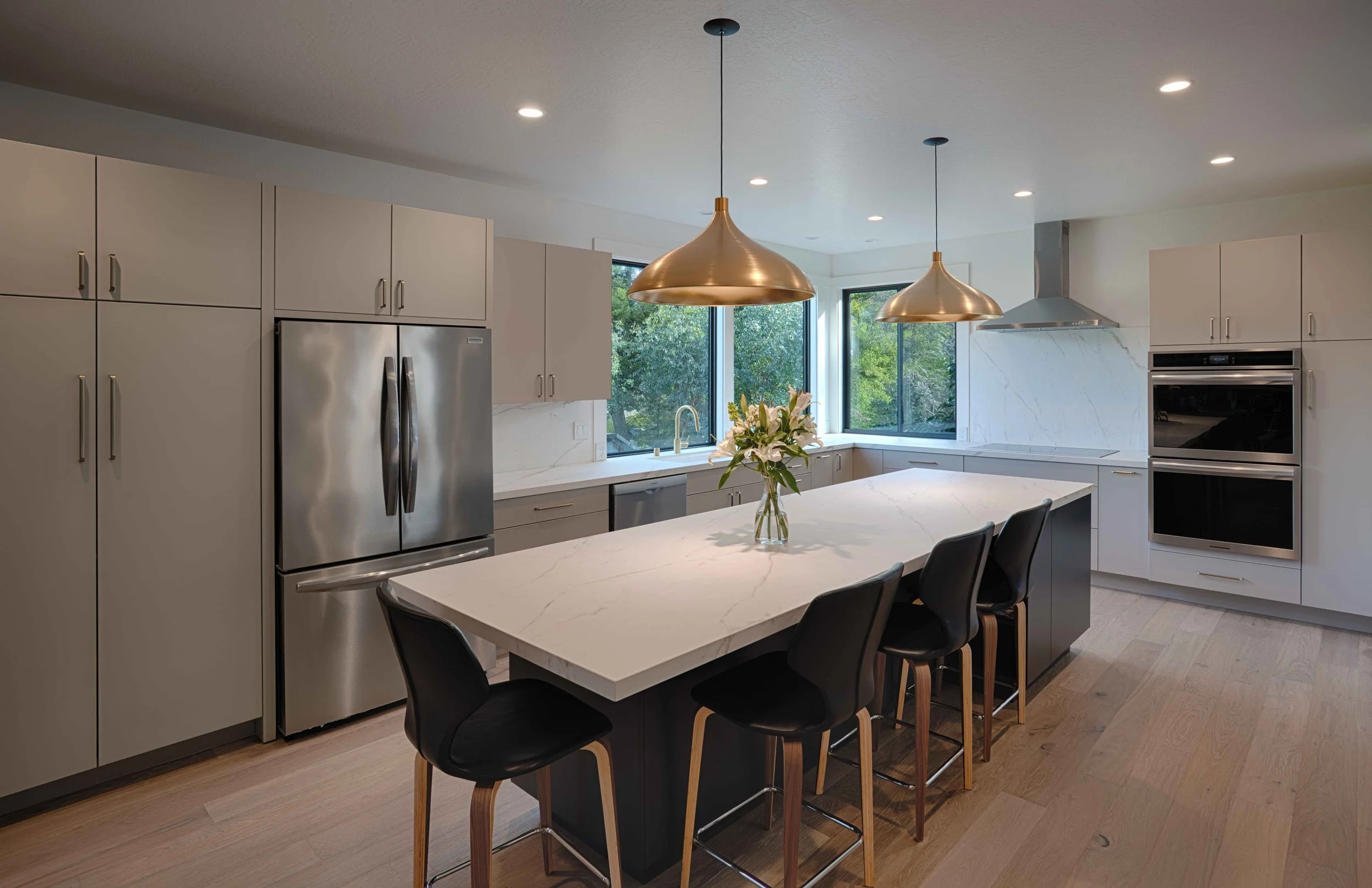 Modern kitchen with large island, black chairs, stainless steel refrigerator, double oven, and pendant lights, with large windows overlooking greenery.