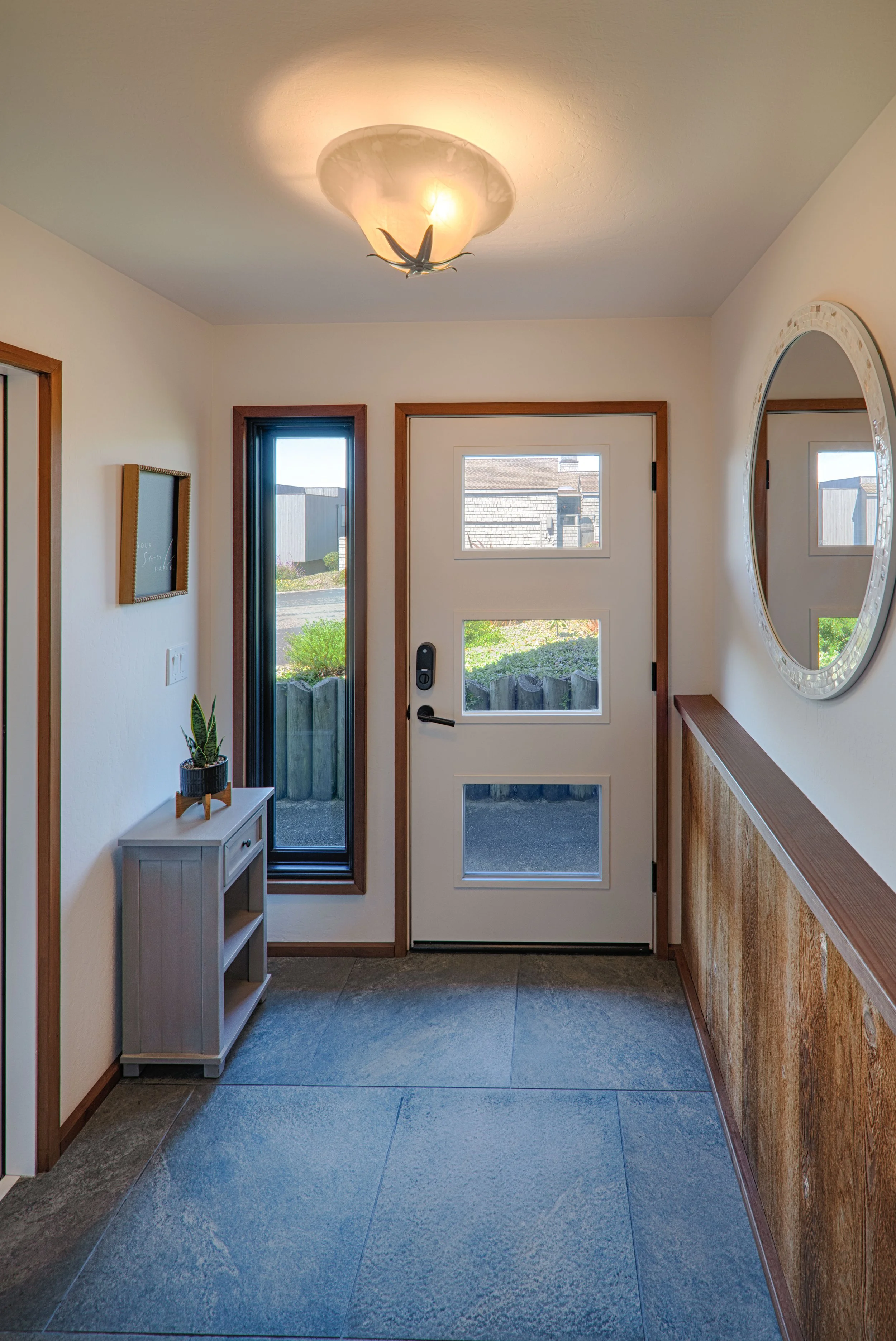 Entryway with front door, small gray table with a plant, narrow window, wall art, and a large mirror on the wall.