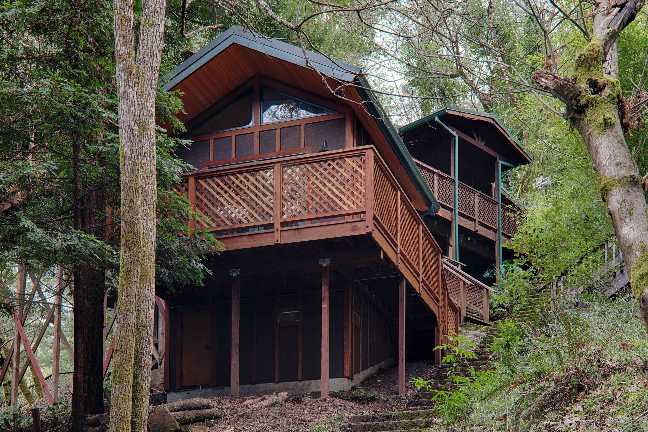 A two-story wooden house built on a hillside surrounded by trees and greenery with multiple decks and stairs.