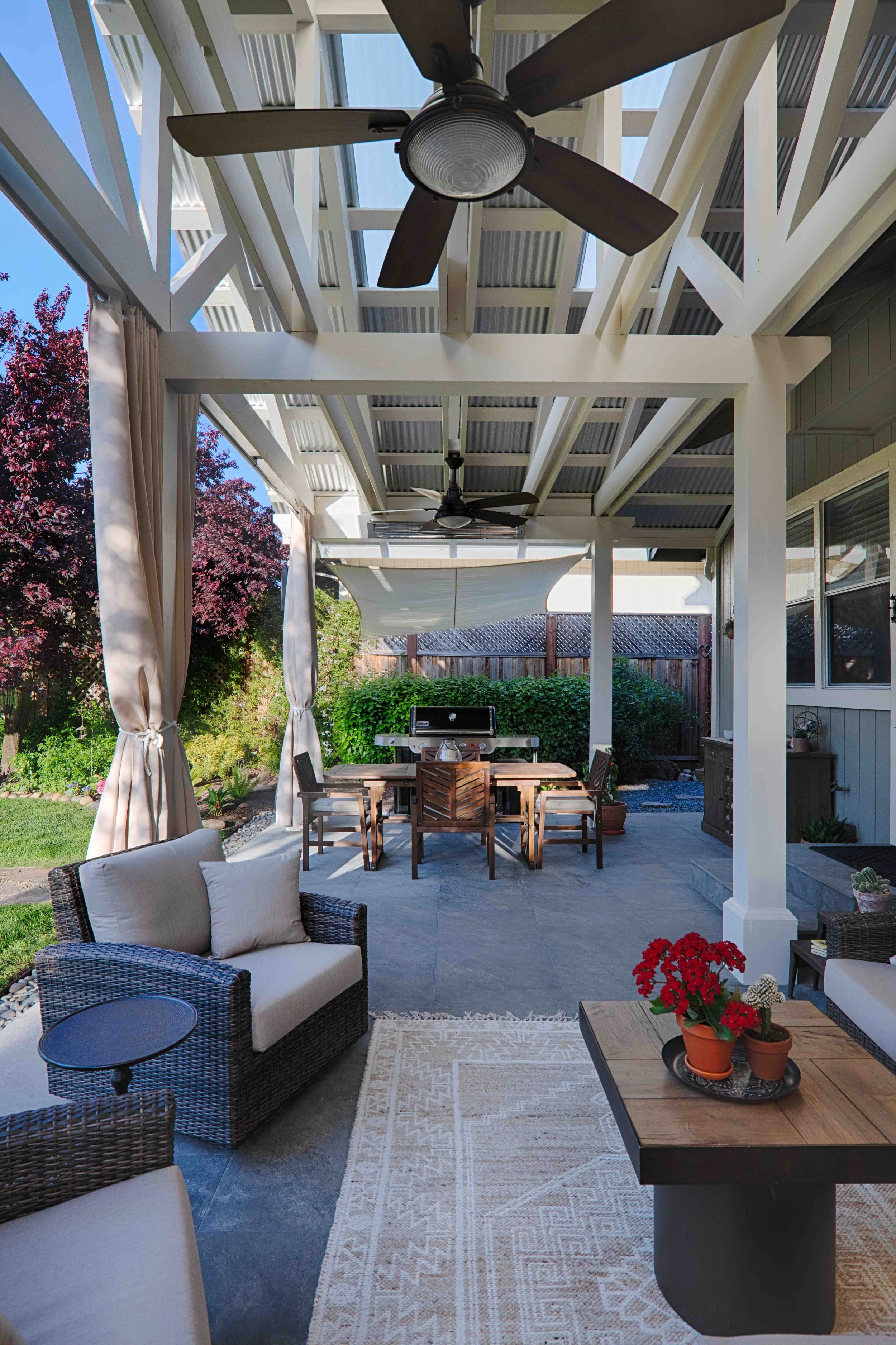 Covered backyard patio with seating area featuring wicker chairs and cushions, a wooden table with potted red flowers, a dining table with matching chairs, a grill, ceiling fans, and outdoor curtains, with garden trees and shrubs in the background.