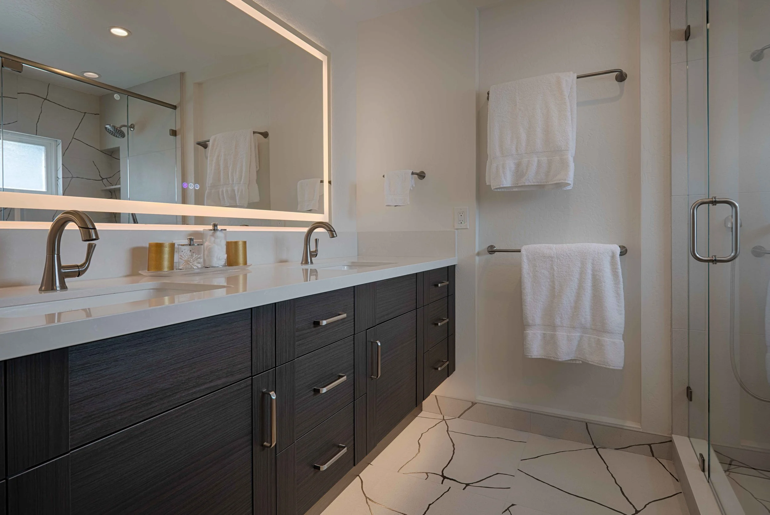 Modern bathroom with double vanity, large mirror with lighting, white countertop, black cabinets, and folded white towels on towel bars, with a glass shower enclosure.
