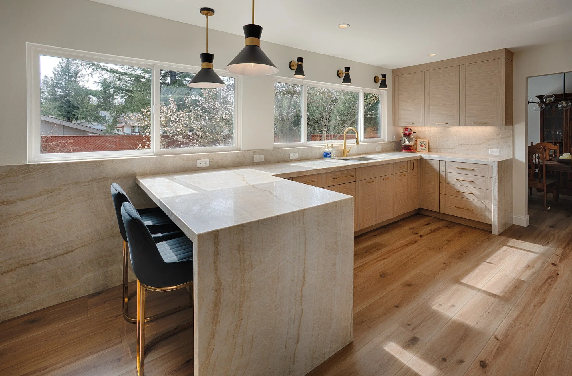 Modern kitchen with light wood cabinets, marble countertops, large windows, black and gold pendant lights, and a view of trees outside.