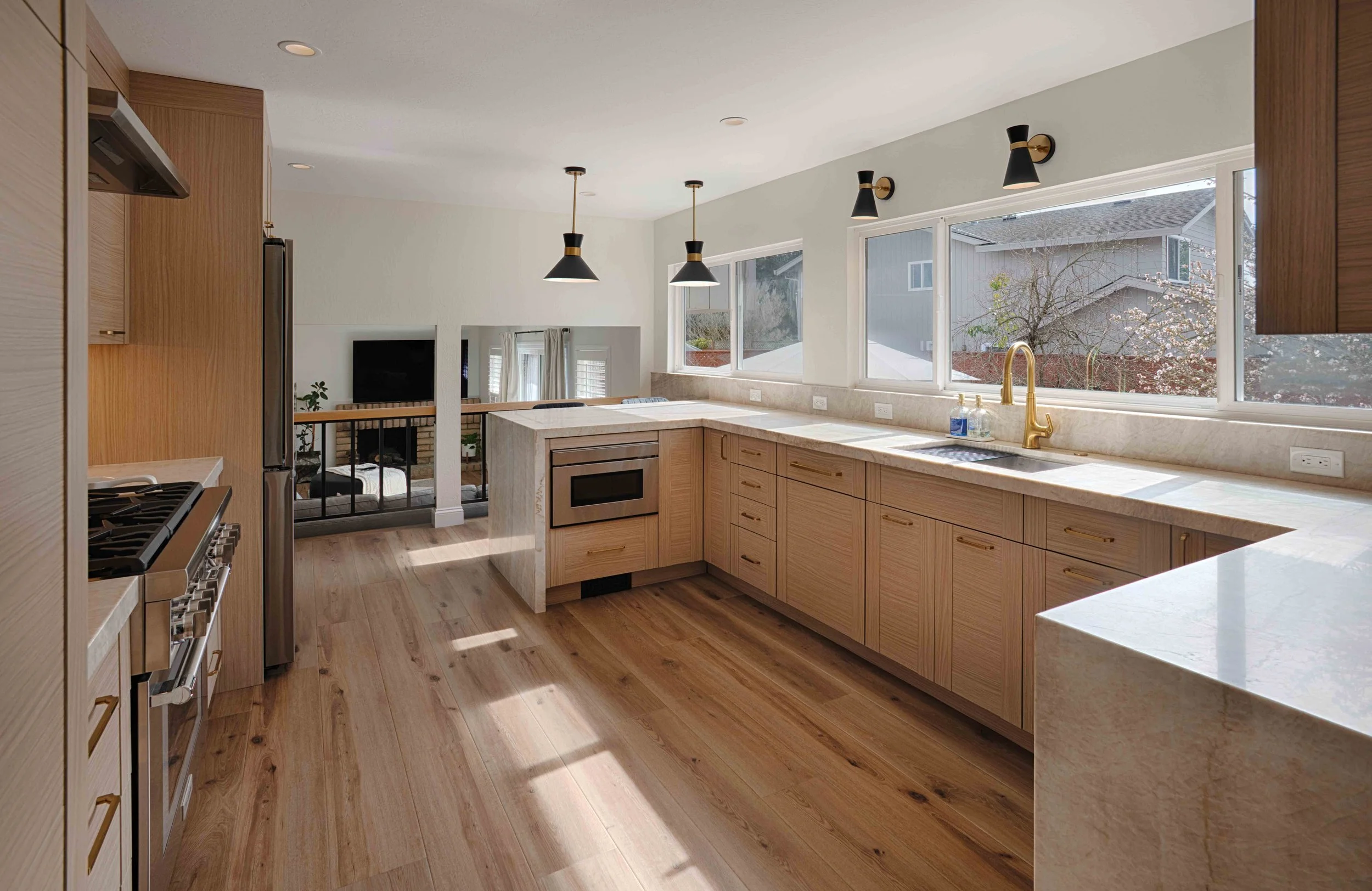 Modern kitchen with light wood cabinets, marble countertops, and large windows letting in natural light. There are black pendant lights hanging from the ceiling and a view into the living area with a TV and outdoor scenery visible through other large