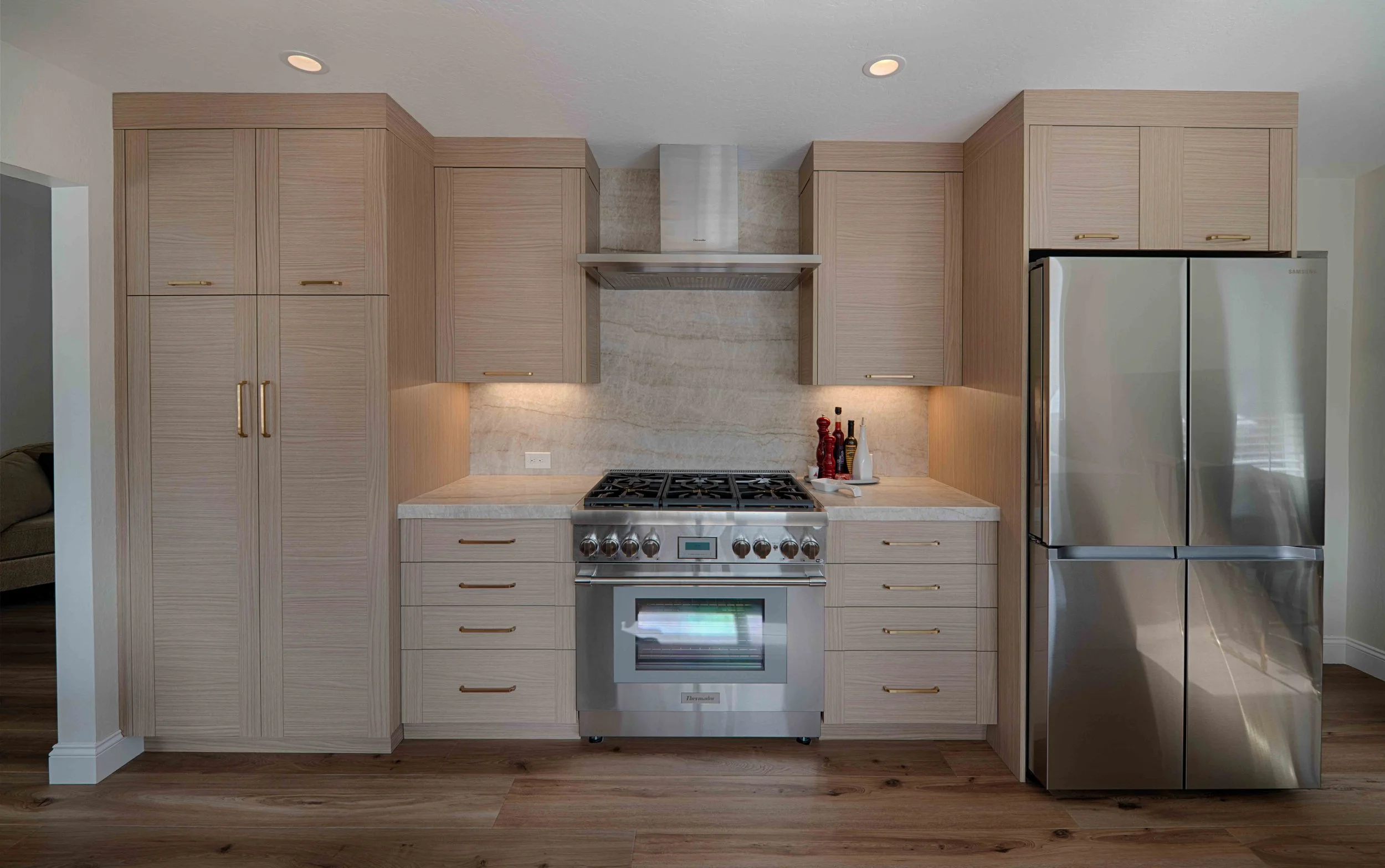 Modern kitchen with light wood cabinets, stainless steel stove, and a refrigerator, with a marble backsplash and hardwood floors.