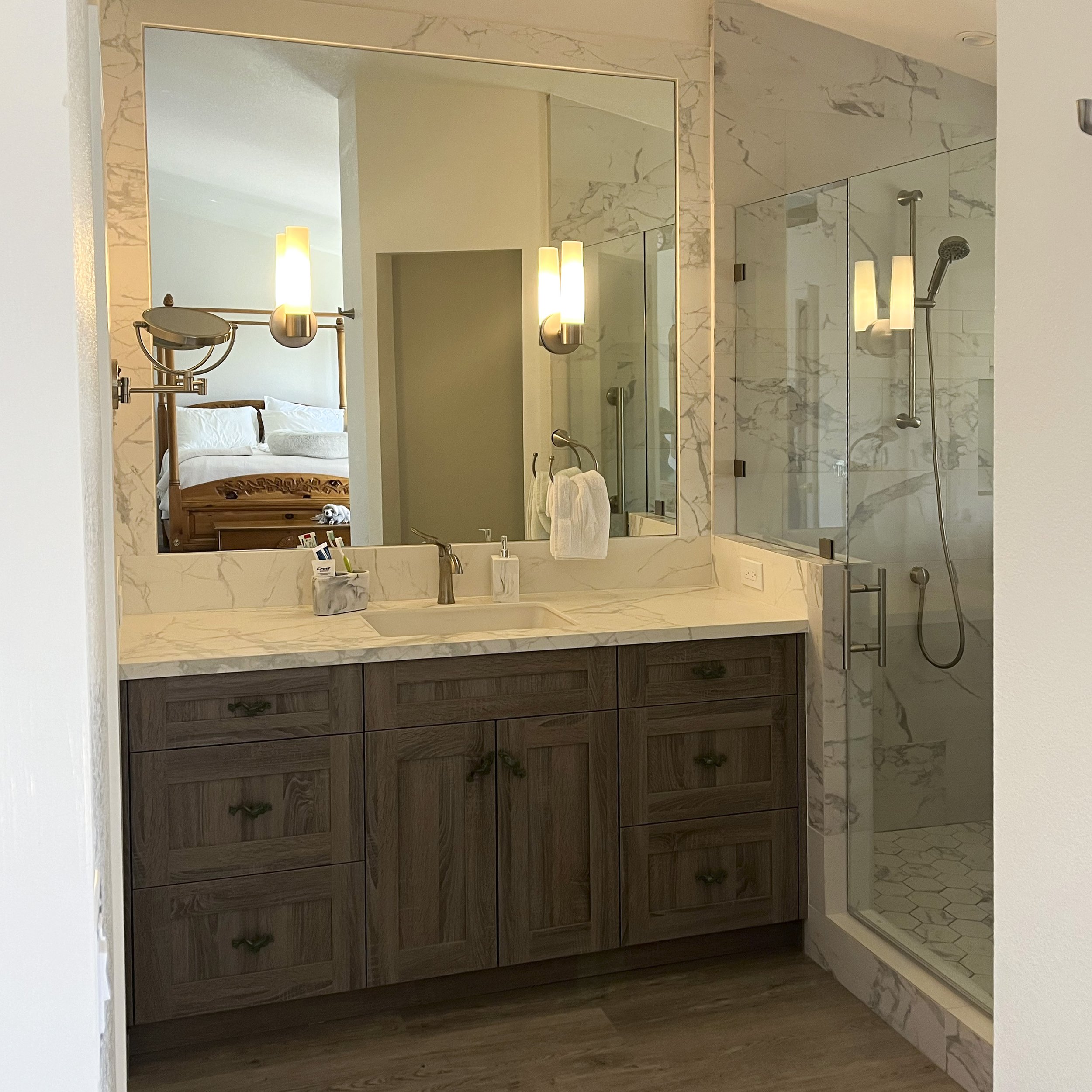 A bathroom vanity with a marble countertop, a sink, and a mirror. There are two wall-mounted lights on either side of the mirror. The shower area with glass walls and a showerhead is visible on the right. A towel hangs on a hook beside the mirror, and a toothbrush holder and small container are on the counter. In the reflection of the mirror, a bedroom with a bed and wooden headboard is visible.