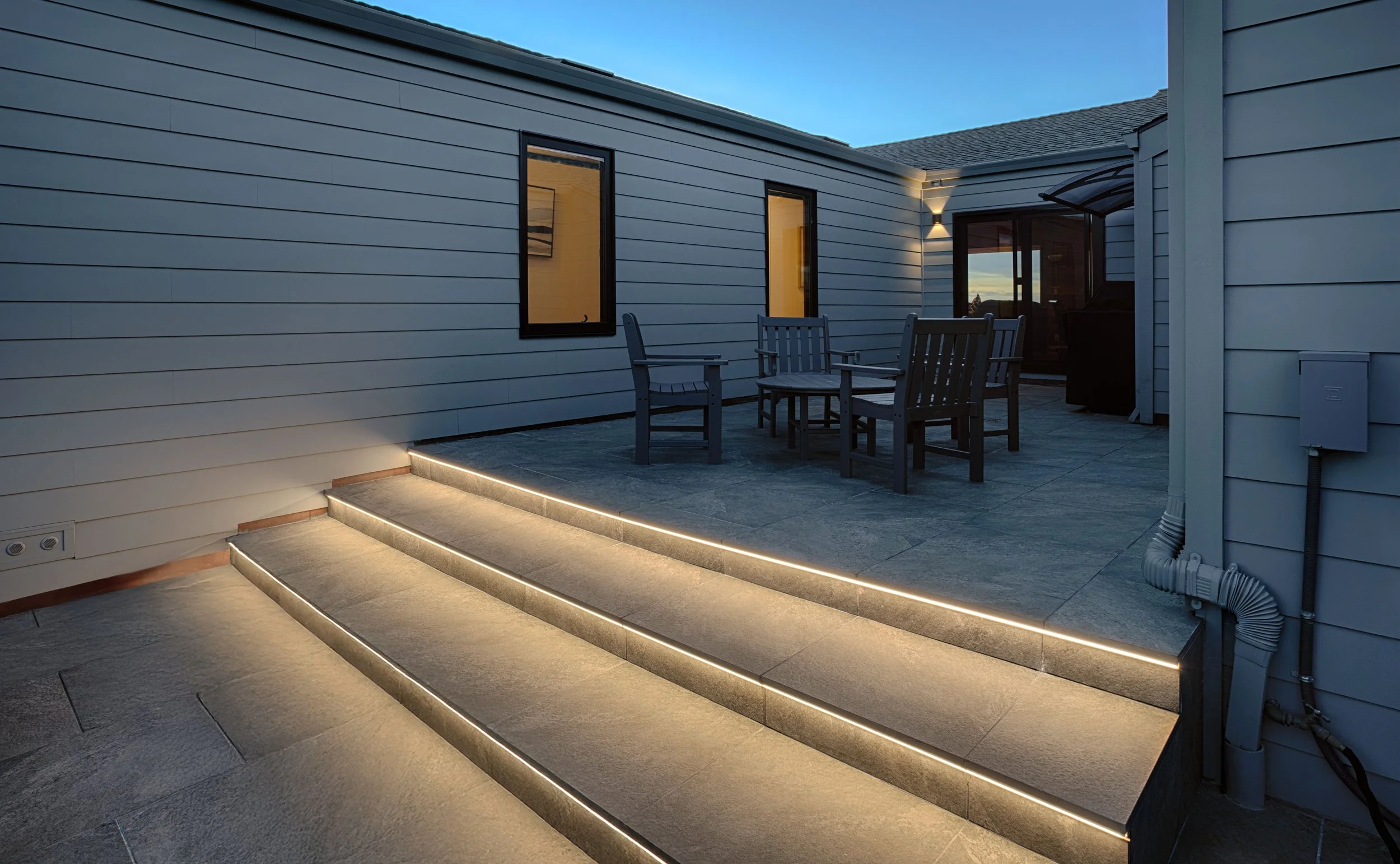 Outdoor patio with steps illuminated by LED lighting, wooden table with four chairs, house siding, windows, and sliding glass door at dusk.
