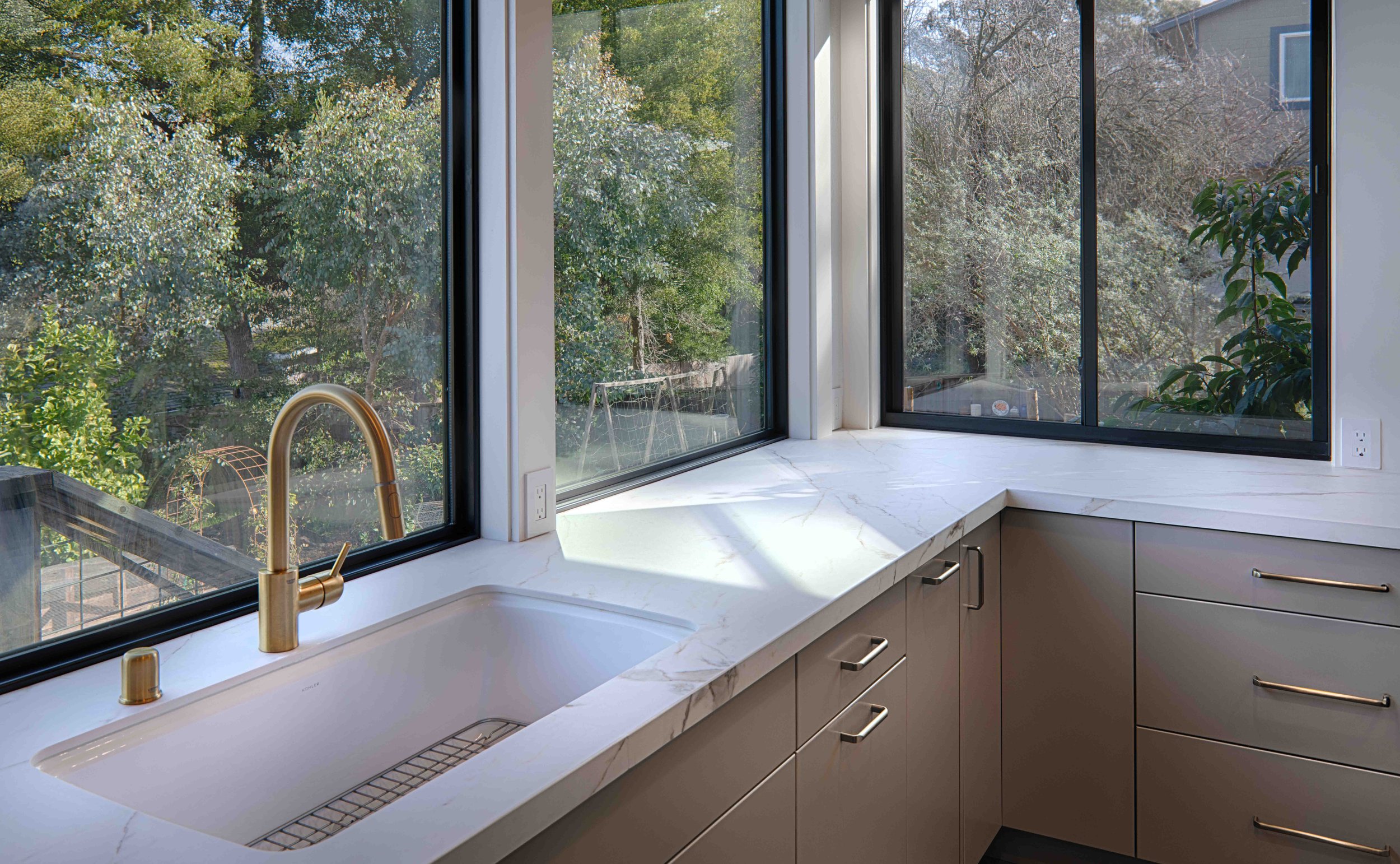 Modern kitchen with large windows overlooking a lush, green outdoor space, beige cabinets, a white marble countertop, and a gold faucet.