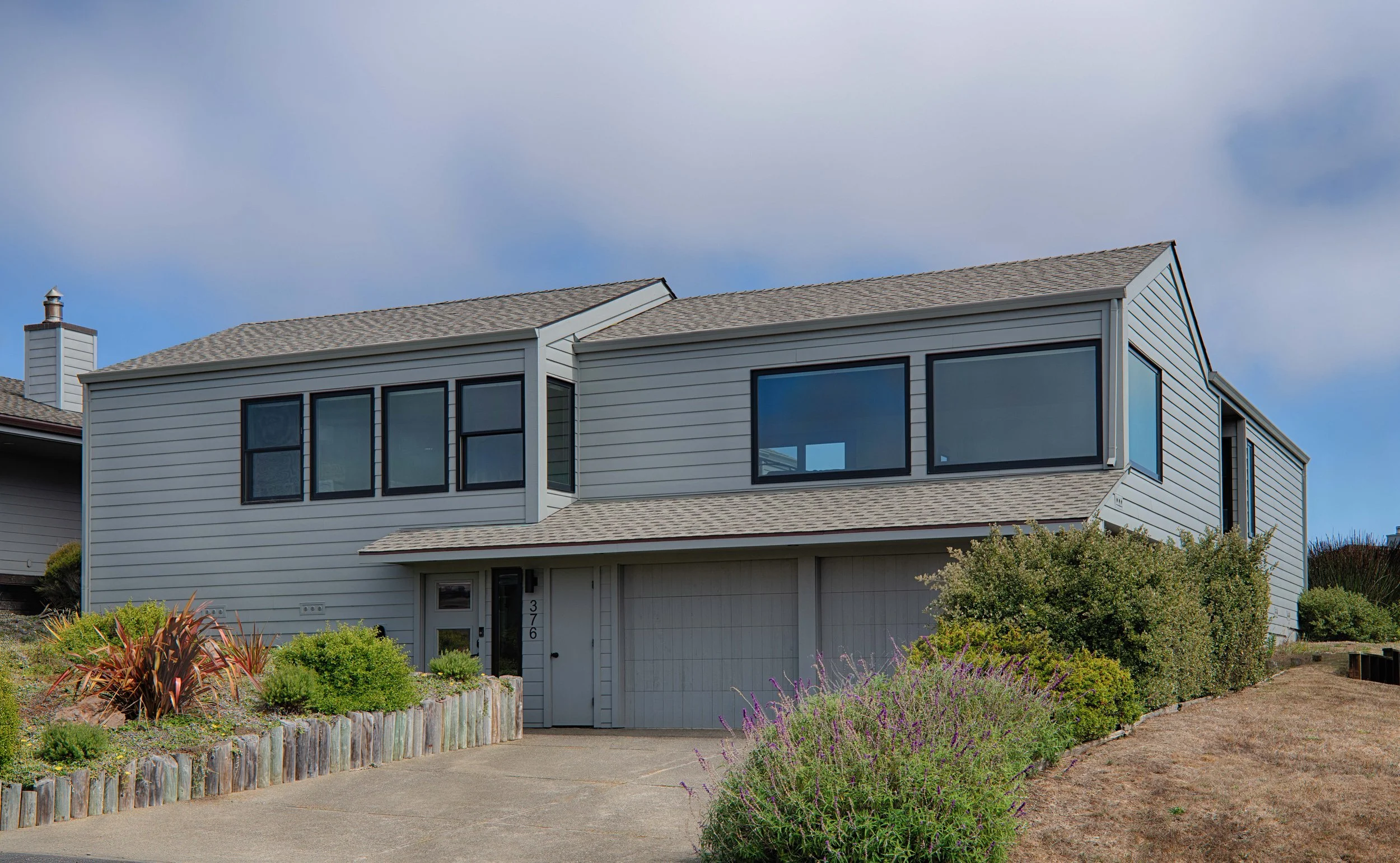 A two-story modern house with gray siding and large black-framed windows, surrounded by landscaped garden with shrubs and purple flowers, and a concrete driveway leading to a garage.