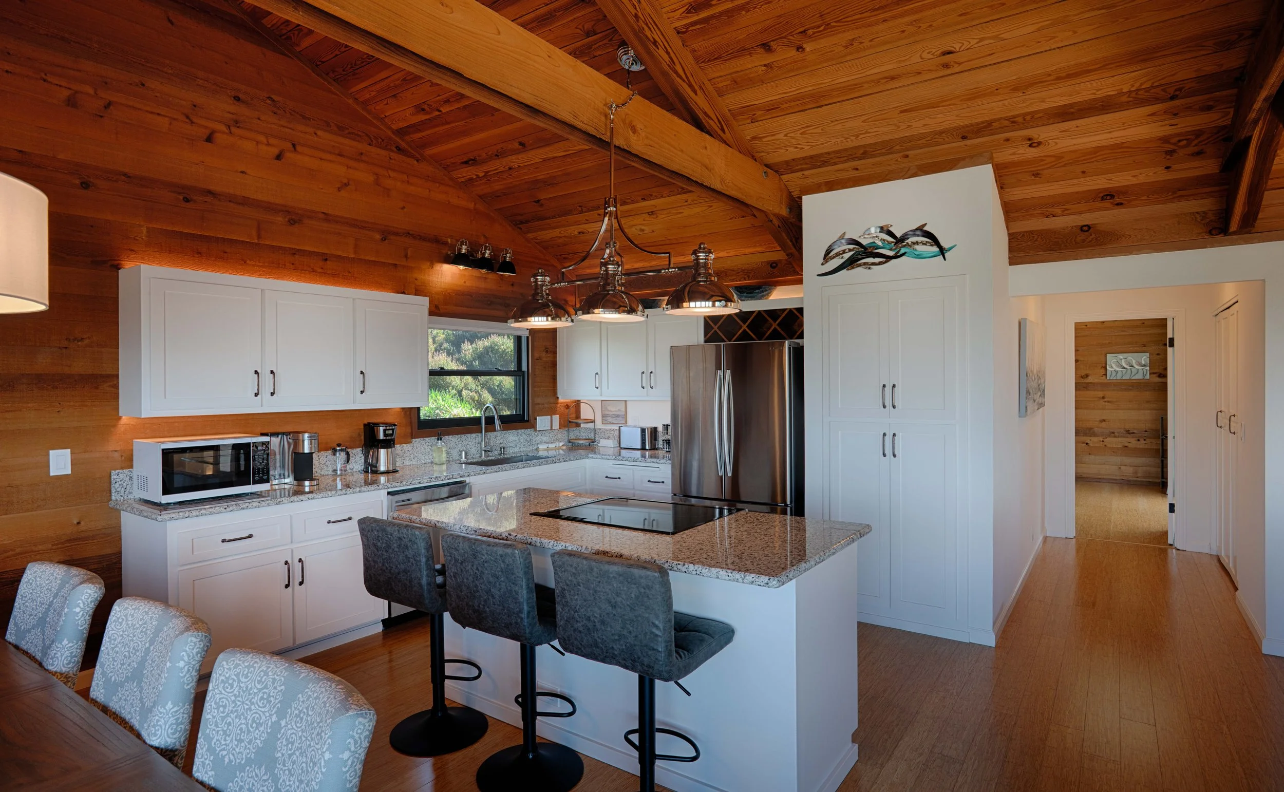 A modern kitchen with white cabinets, granite countertops, and a stainless steel refrigerator. The room features a wooden ceiling with exposed beams, a window over the sink, a microwave, coffee maker, and bar stools at a kitchen island.