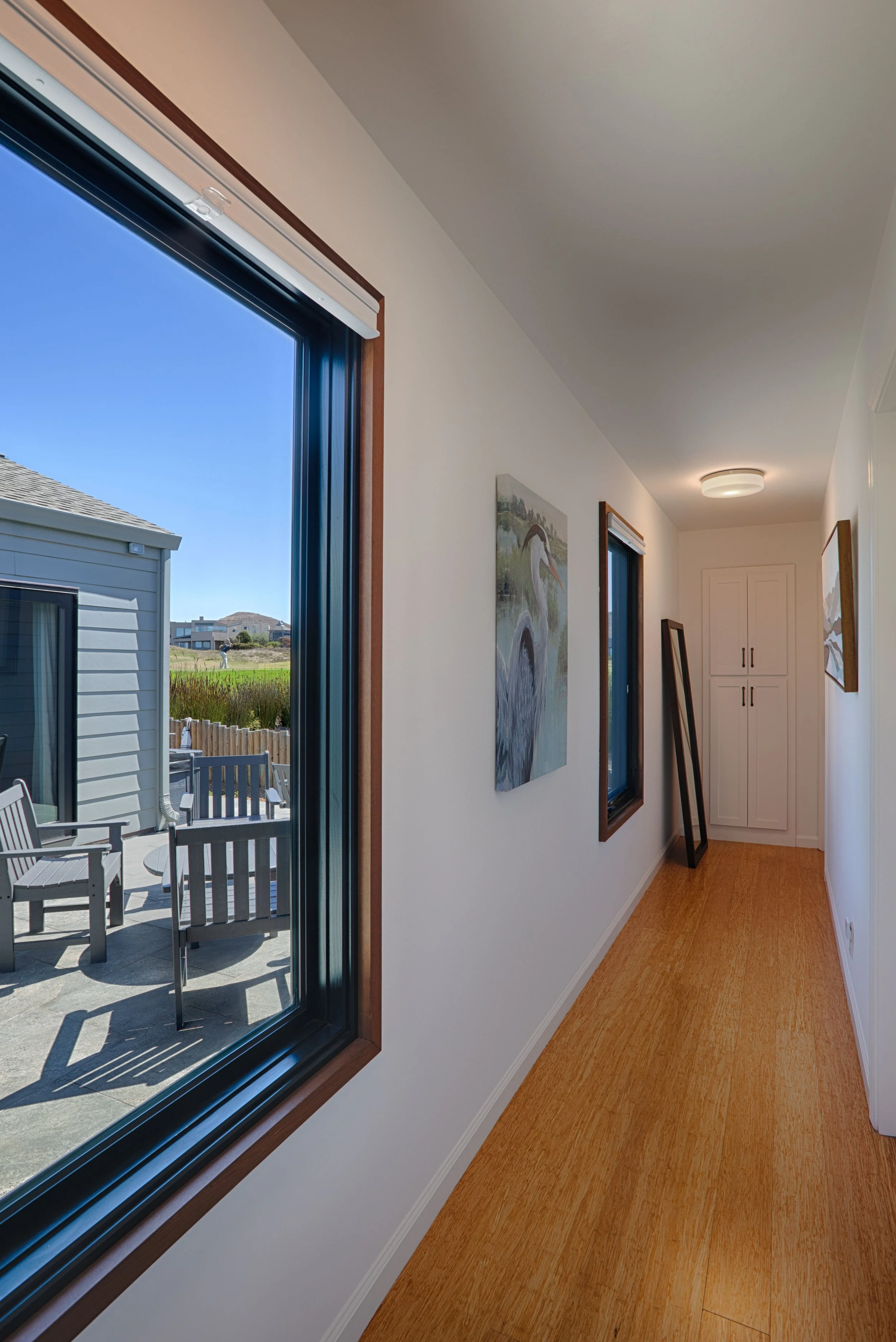 A hallway with wooden flooring and white walls, decorated with paintings, leading to a white cabinet at the end. There are two windows with dark frames, and outside, a patio with chairs and a view of neighboring houses and a grassy field. Sunlight is