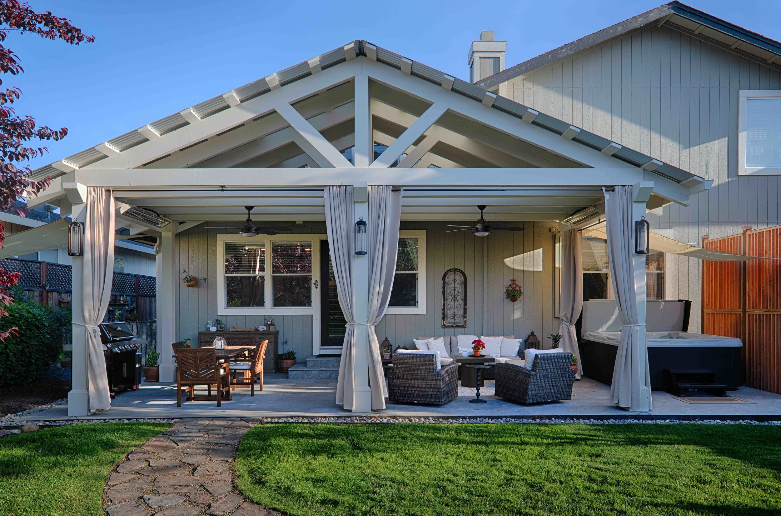 Backyard patio with outdoor seating, dining area, hot tub, and decorative lighting under a white pergola, adjacent to a house with gray siding and a stone pathway leading to green grass.