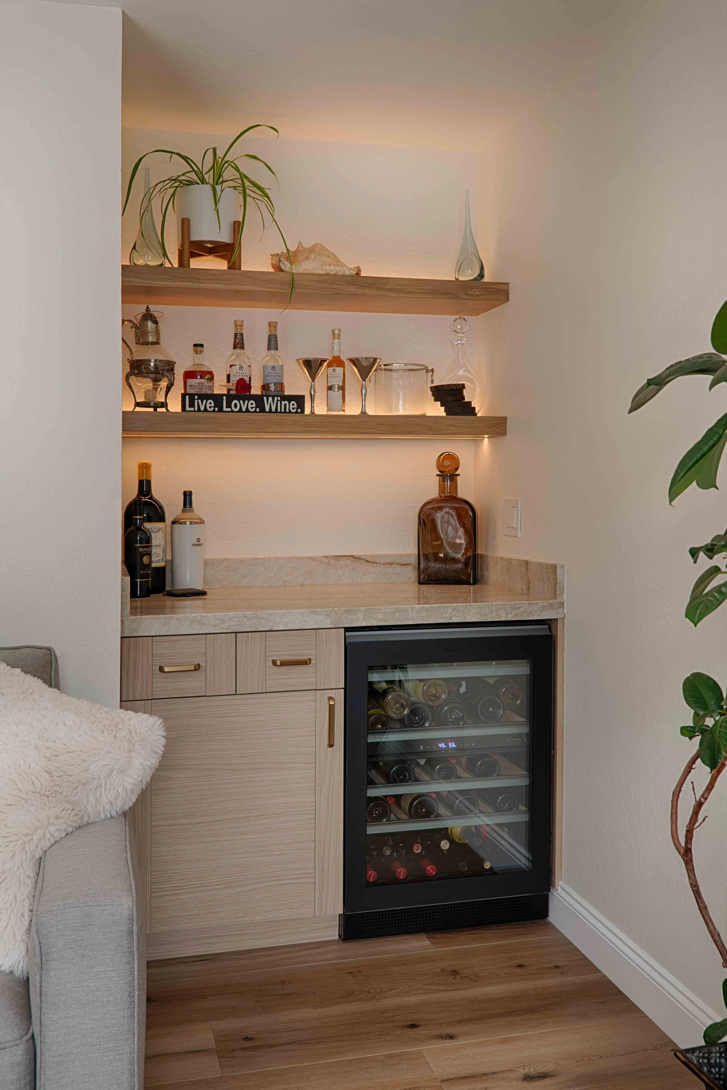 A home wet bar with two wooden shelves holding various bottles, glassware, and decorative items, alongside a counter with a few bottles and a wine cooler below.