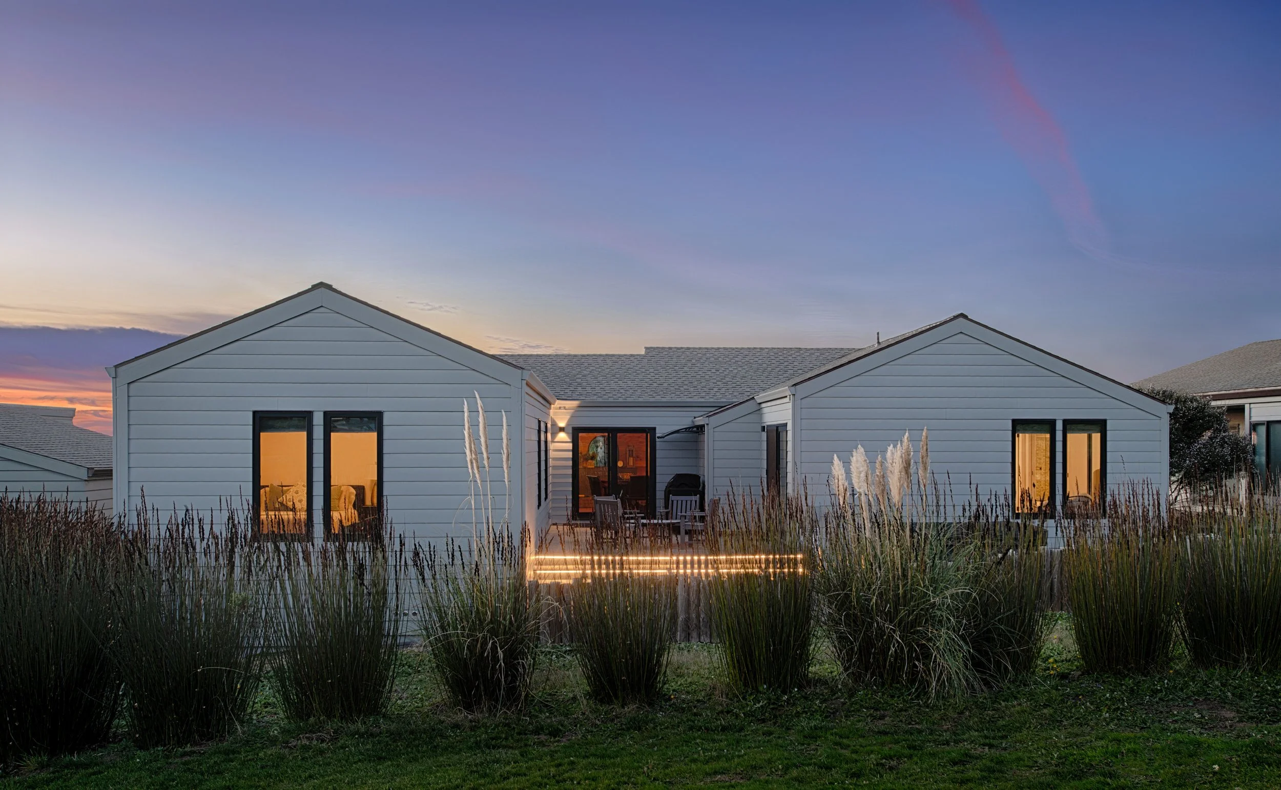 A modern two-story house with white horizontal siding, large windows, a backyard deck with string lights, surrounded by tall grasses, during sunset or dusk.