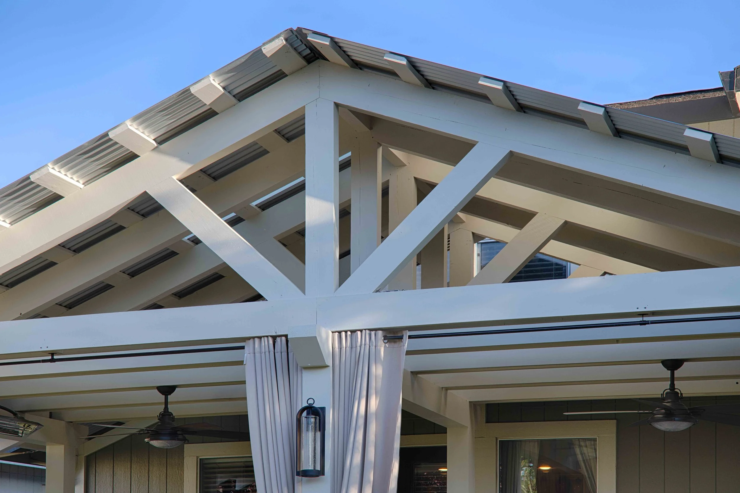 Close-up of a modern house's roof structure with white painted wooden beams, metal roofing sheets, and a decorative outdoor lampshade, under a clear blue sky.