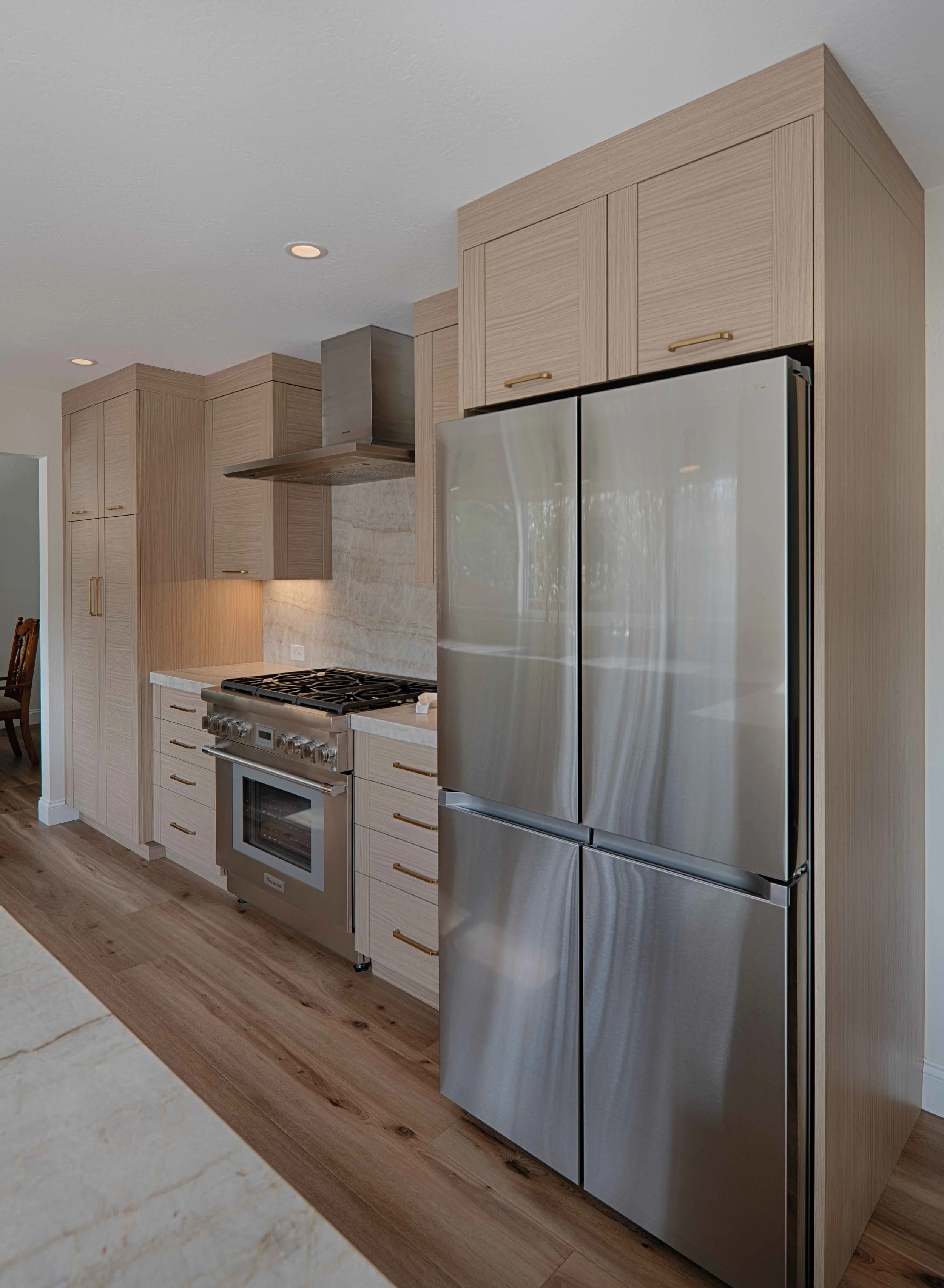 Modern kitchen with light wooden cabinets, stainless steel refrigerator, stove, oven, and marble backsplash, with overhead recessed lighting.