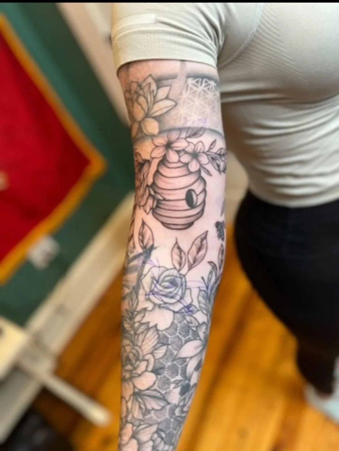 Close-up of a person's arm with a detailed black and gray tattoo featuring flowers, a honey jar with a bee, and honeycomb patterns.