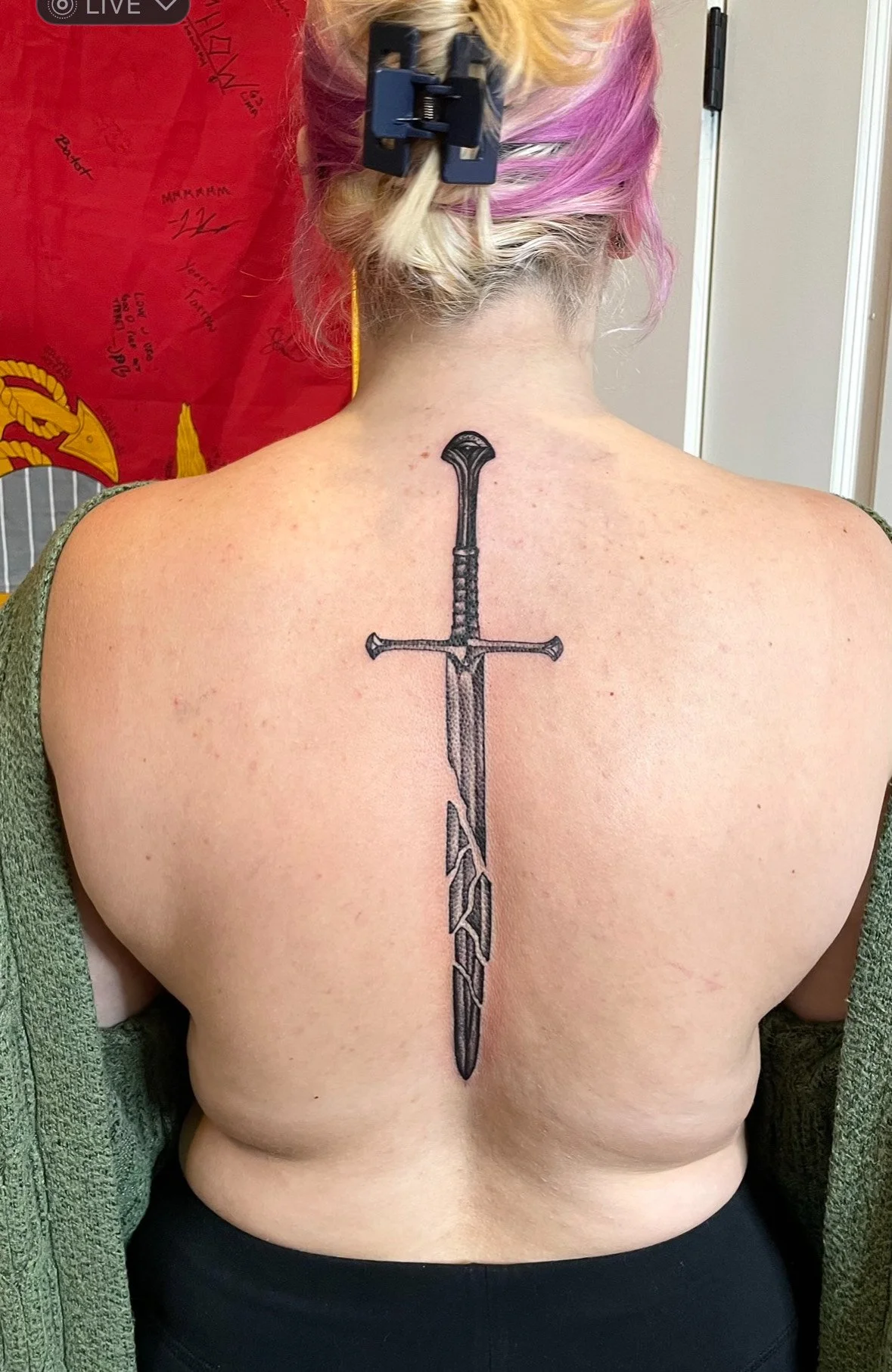A woman with colorful pink and blonde hair getting a tattoo of a sword on her upper back.
