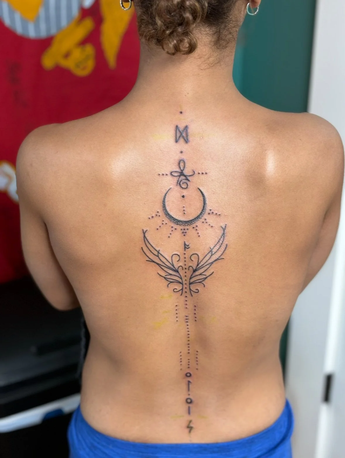 Back view of a person with a geometric and celestial tattoo running along the spine, featuring various symbols, moons, wings, and lines.