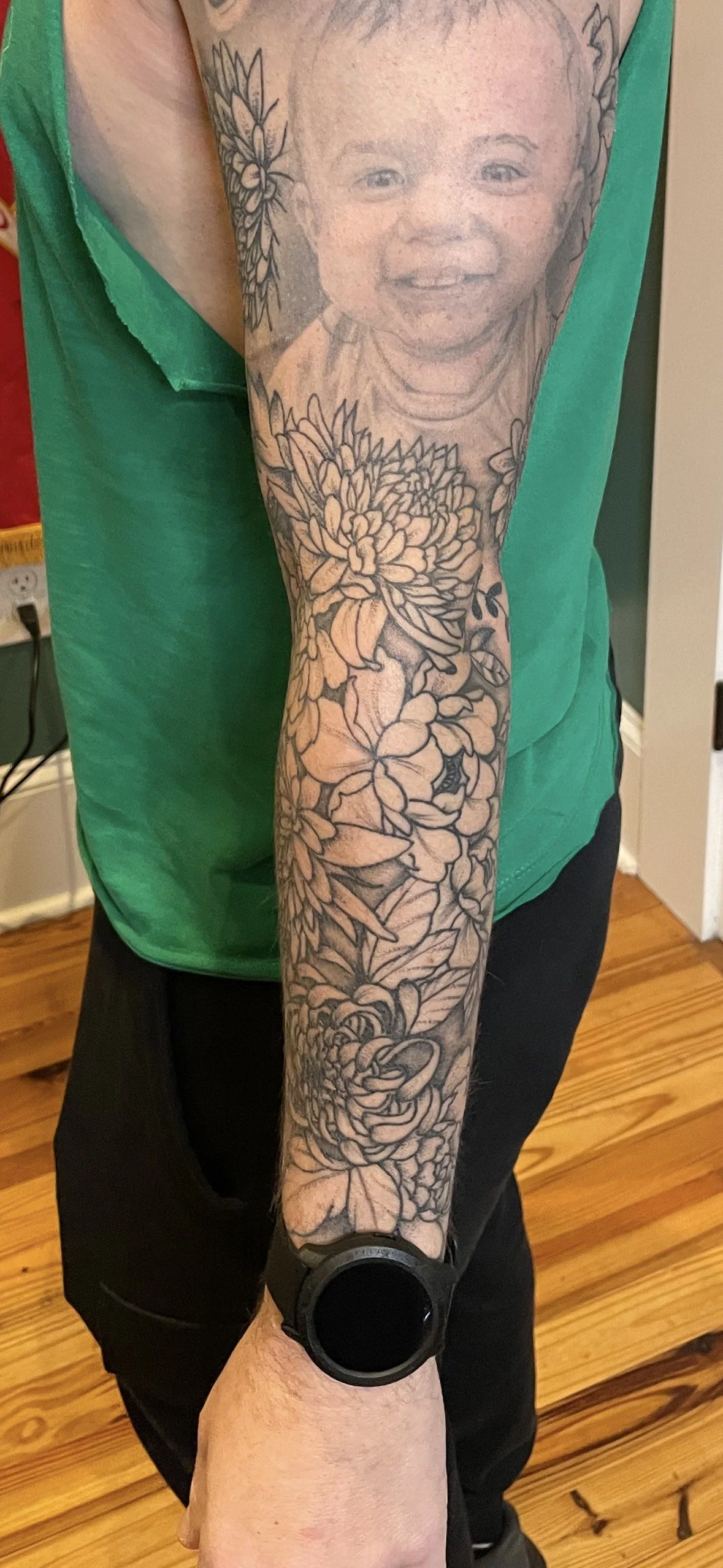 A person's arm with a detailed tattoo of a child's face and flowers extending from the shoulder to the wrist.