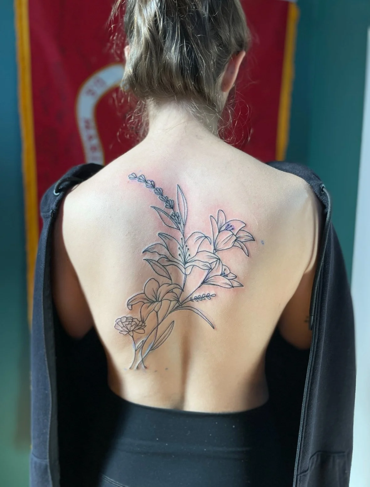 A woman with her back to the camera shows a large, delicate floral tattoo on her back, with outlines of flowers and leaves, partially covered by her open black jacket.