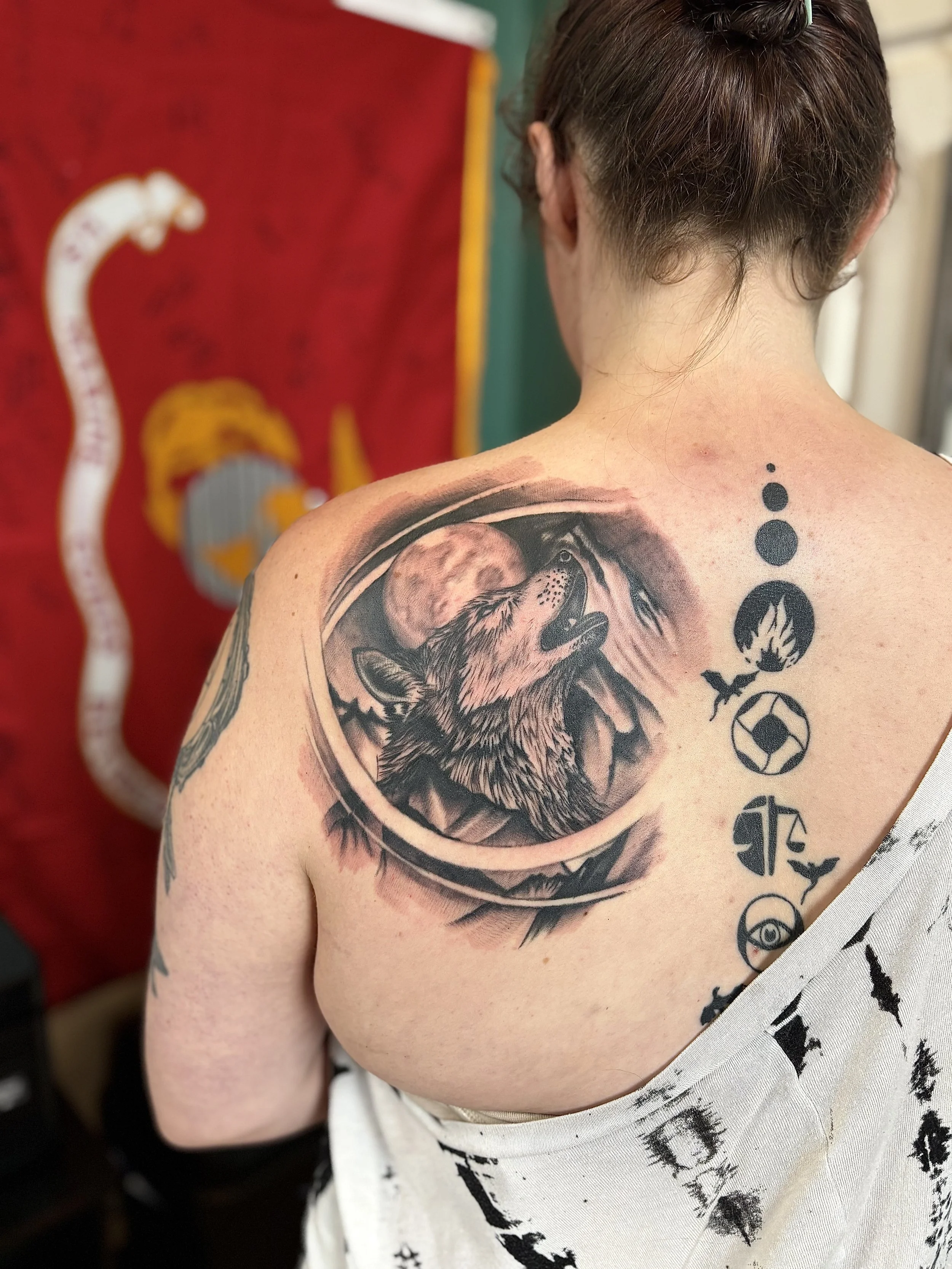 Tattoo of a howling wolf under a full moon on a person's shoulder and upper back, with a series of black symbols running down the back.