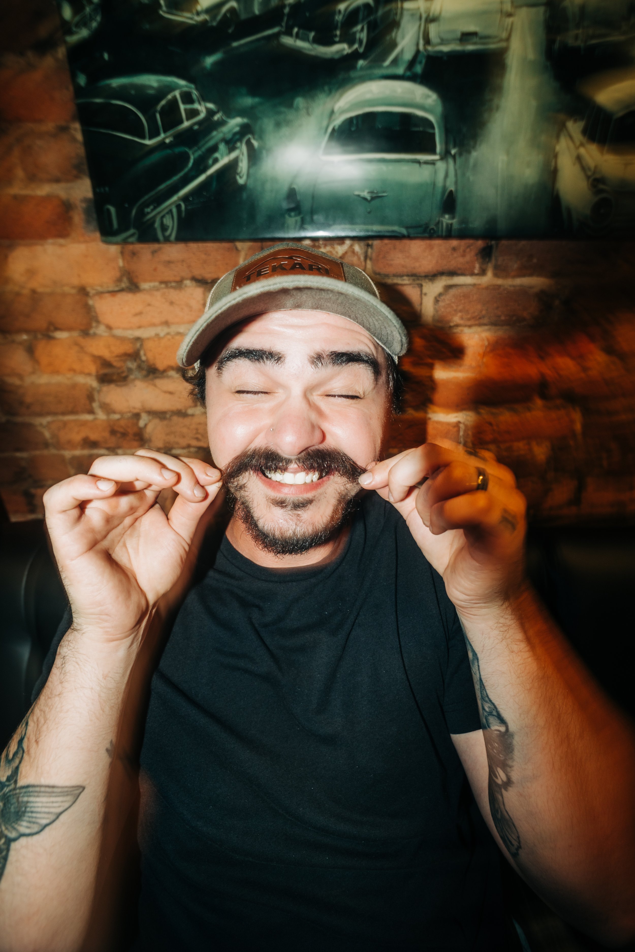 A man with a beard laughing, holding his mustache with both hands, wearing a baseball cap and a black T-shirt, in front of a brick wall with vintage car artwork.